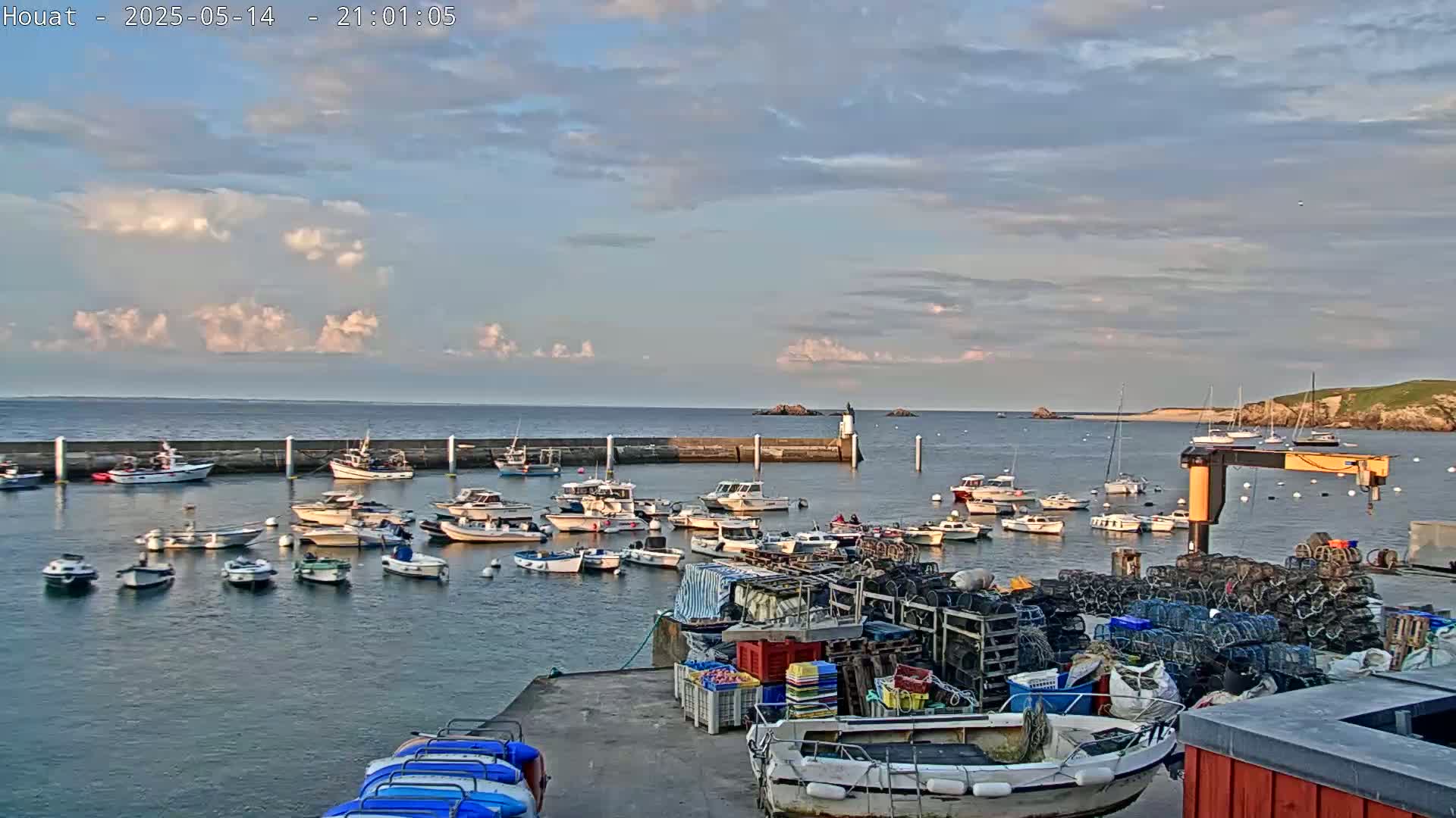 Many small boats are moored in a harbor under a partly cloudy sky at dusk, with a larger body of water and land visible in the background.