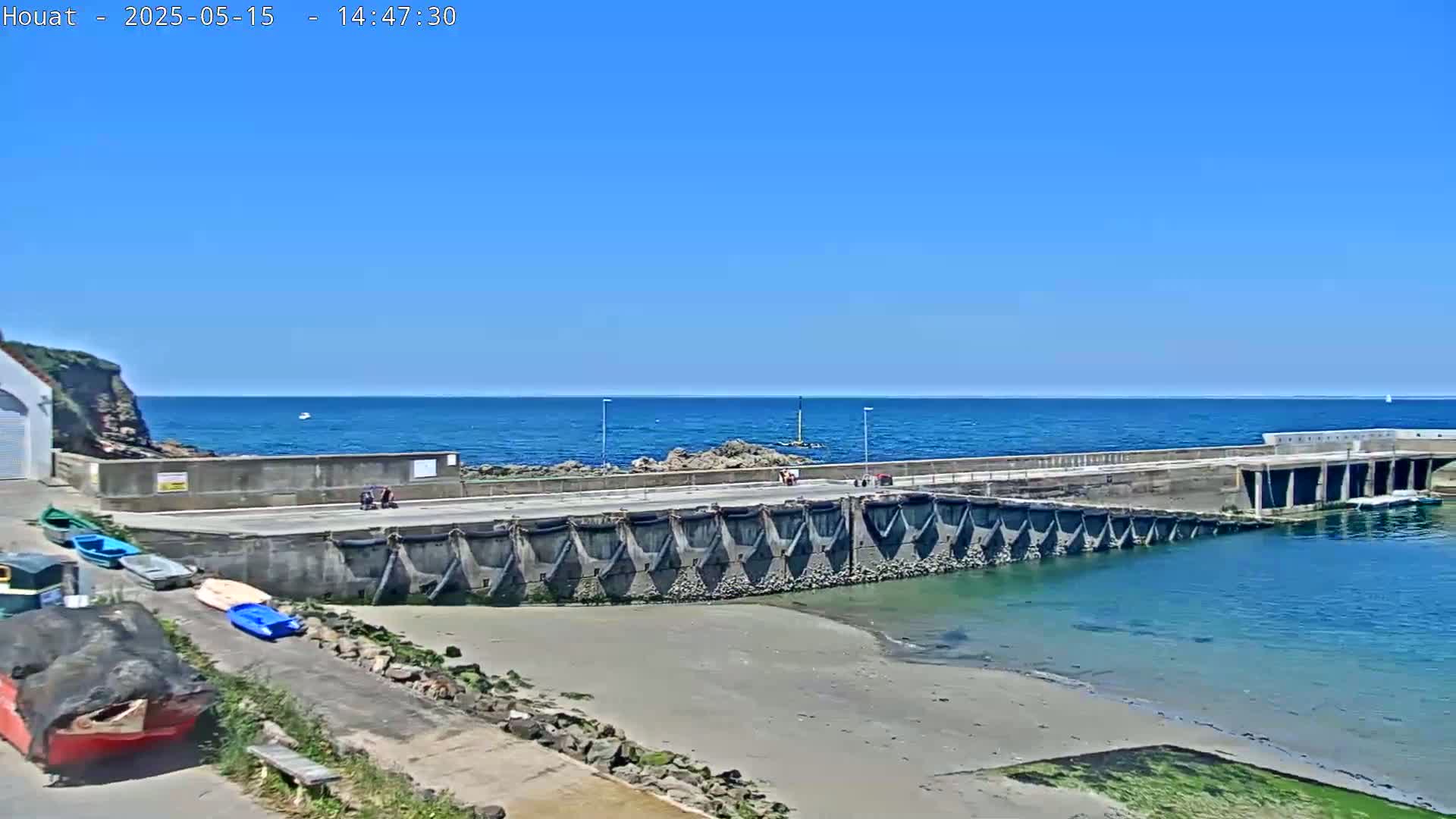 A small harbor with a concrete pier, several small boats, and a sandy beach is visible under a clear blue sky.