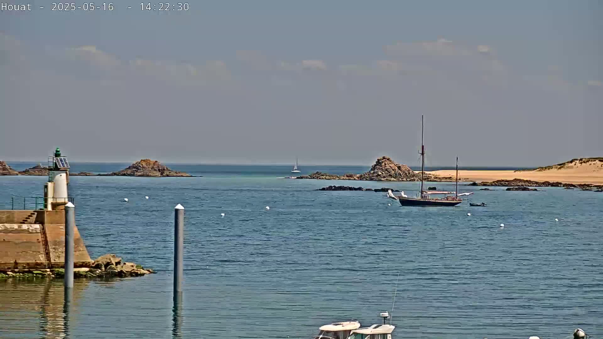 A sailboat and small motorboat are anchored in a calm, sunny bay near a lighthouse and rocky islands.