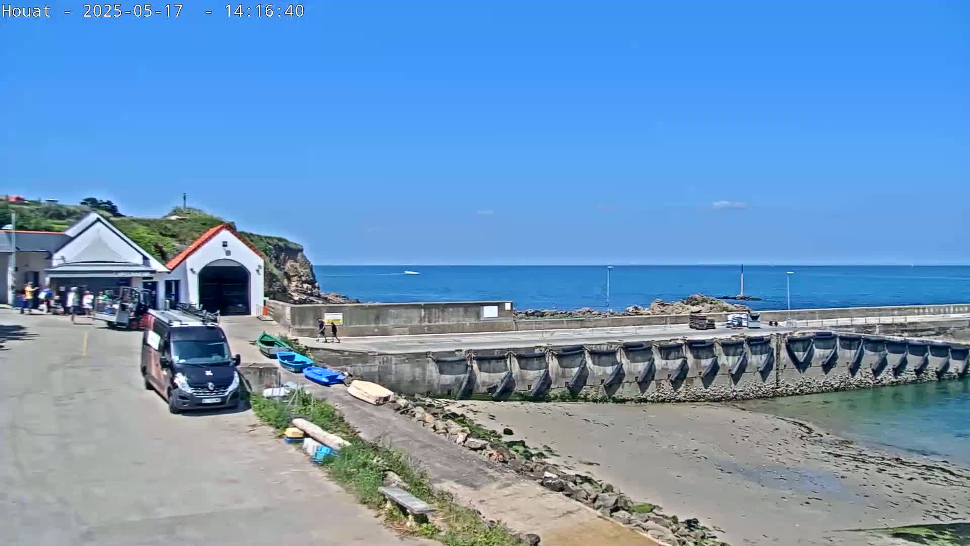 A small harbor with a building, several people, a van, and several small boats is visible on a sunny day with a clear blue sky and calm ocean.