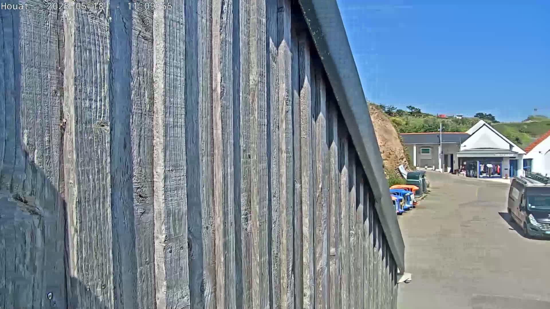 A gray weathered wood fence angles away from the camera toward a sunny parking lot with several small vehicles and buildings.