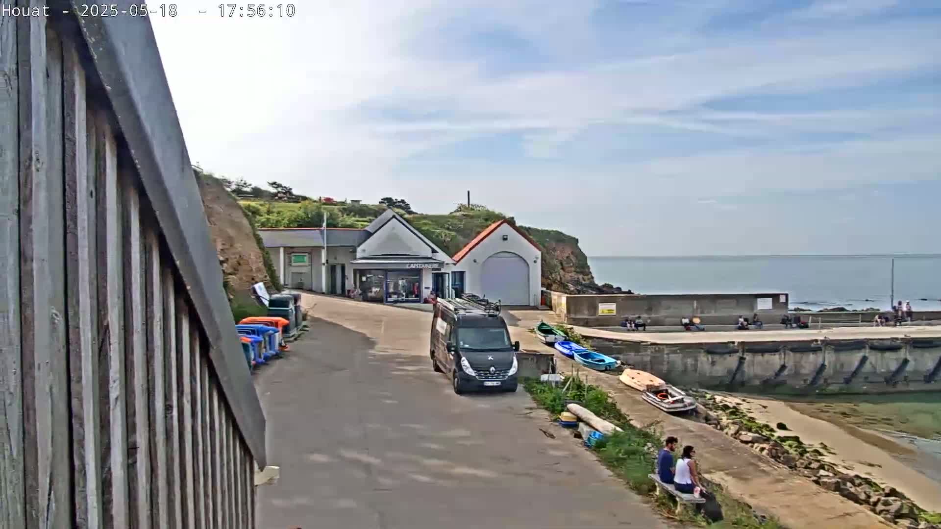 A partly cloudy day at a seaside harbor shows a van parked near a building, several small boats, and a couple sitting on a bench overlooking the calm water.