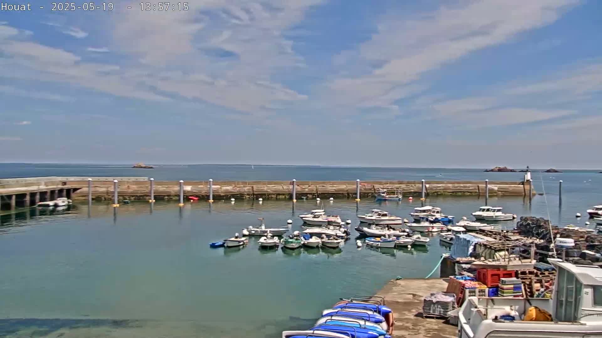 A harbor filled with numerous small boats under a partly cloudy sky, with calm, clear water and a distant rocky shoreline.