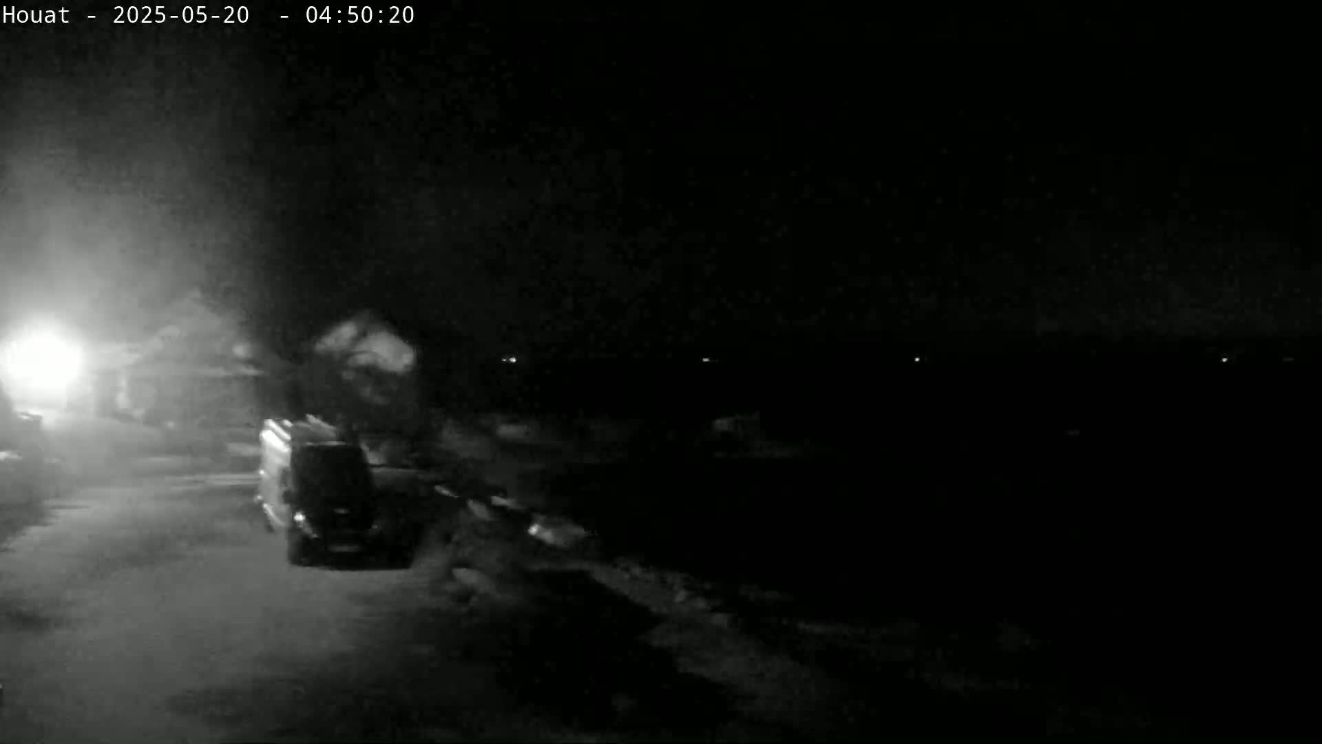 A dark, nighttime scene shows a van parked on a road near some buildings under hazy or foggy conditions.
