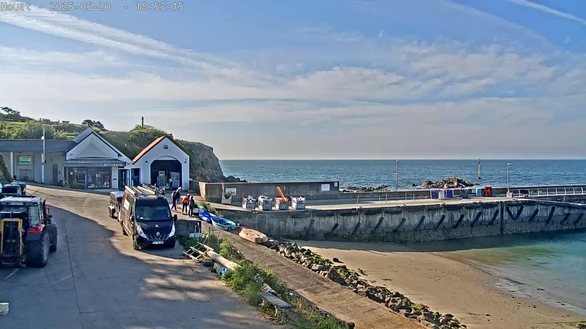 A small harbor with a building, several vehicles, and a few people is shown under a mostly sunny sky with some scattered clouds.
