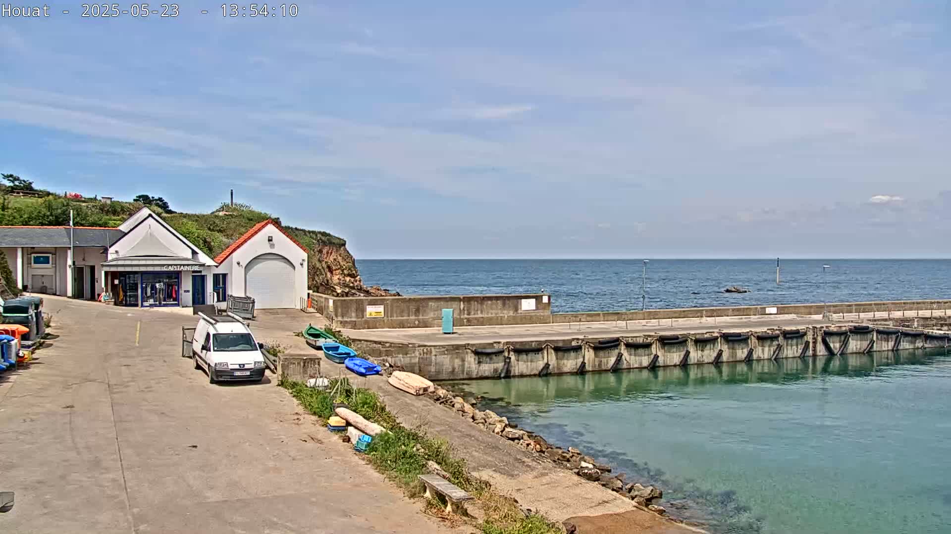 A small harbor with a building and several boats sits beside a calm ocean under a mostly clear sky.
