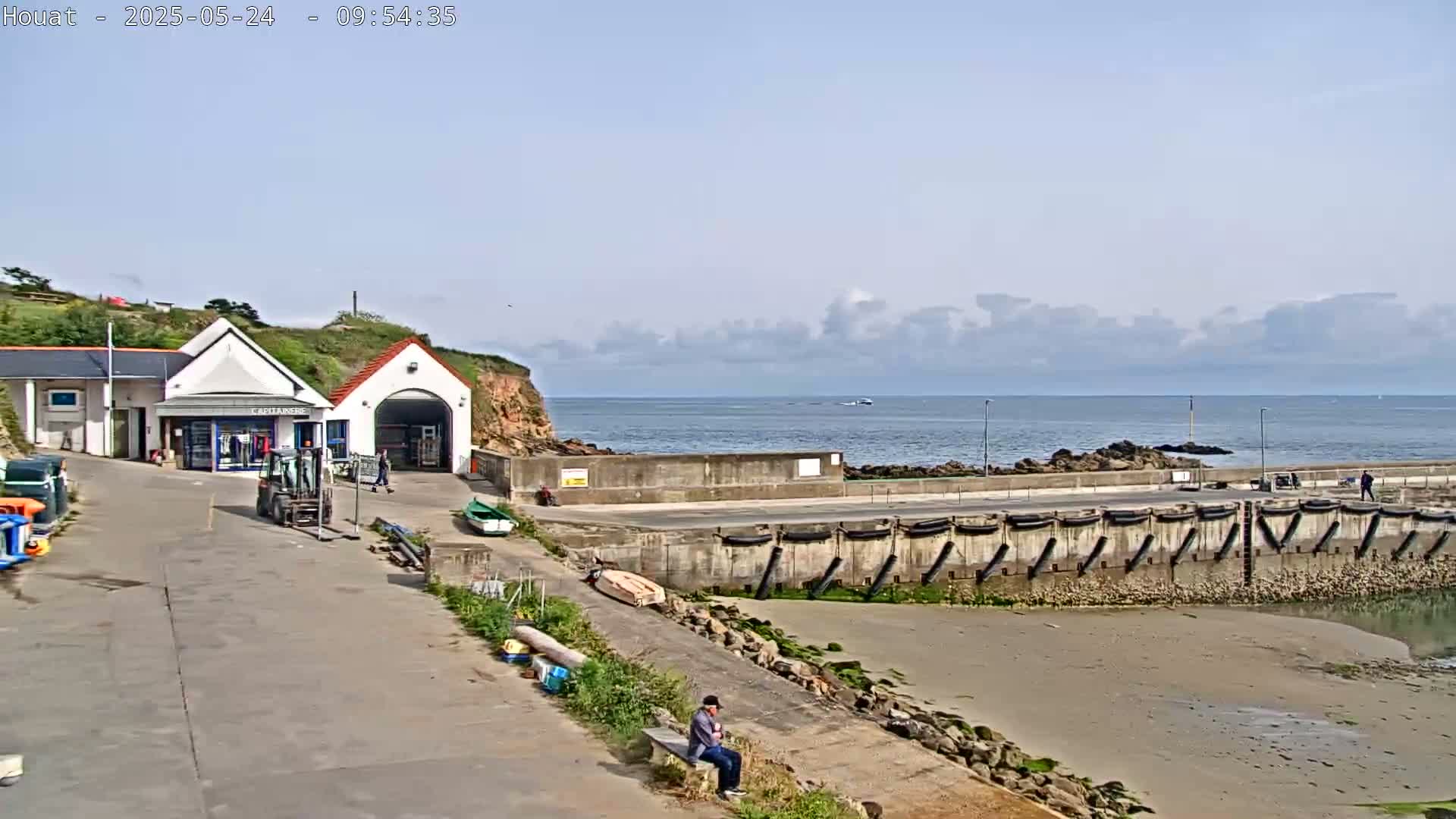 A small harbor with a building and a person sitting on a bench overlooks a calm sea under a partly cloudy sky.