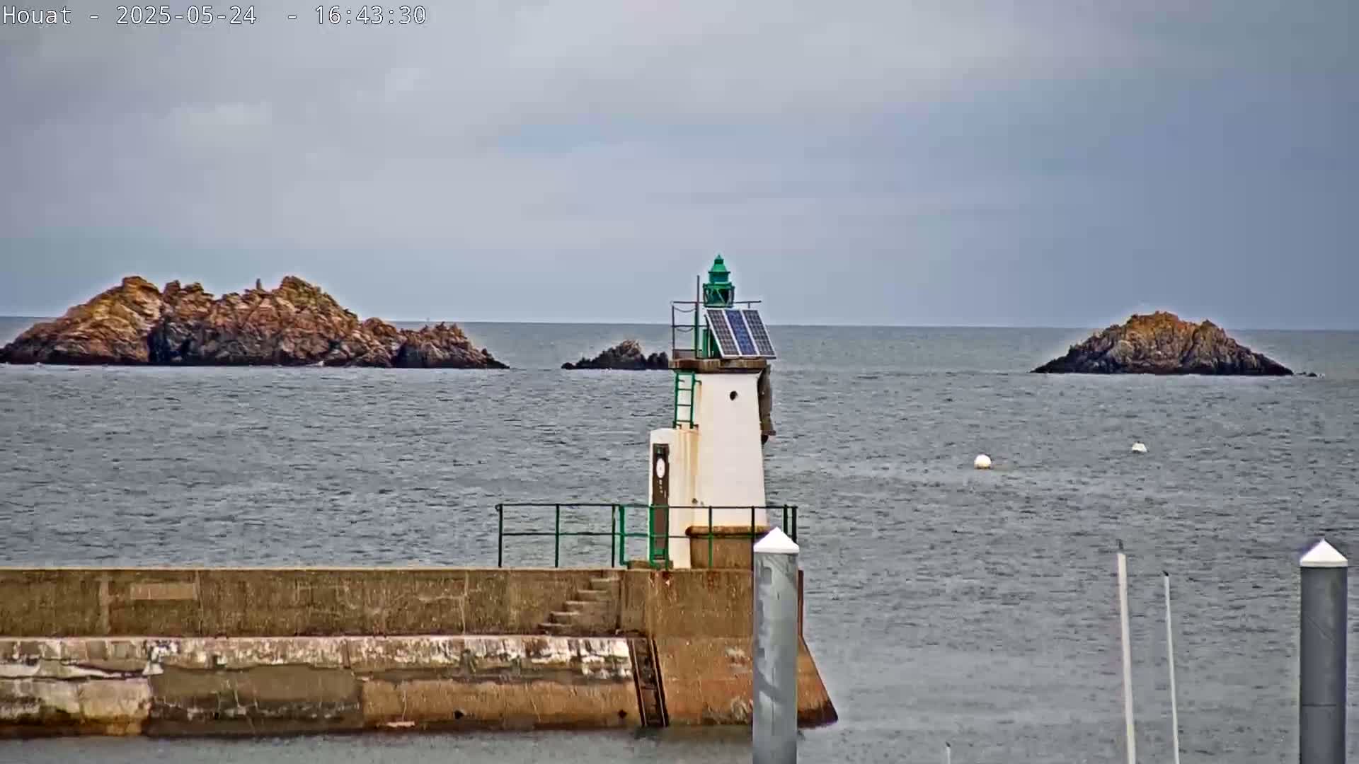 A small lighthouse with a solar panel sits on a seawall overlooking a calm body of water with several small rocky islands in the distance under an overcast sky.
