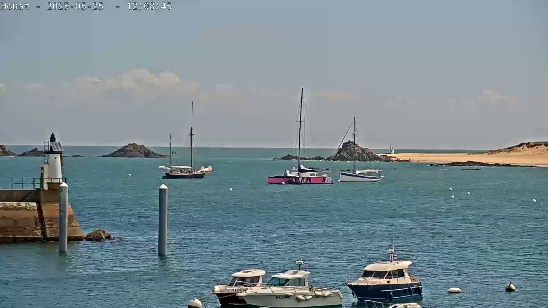 A calm, sunny day shows several sailboats and motorboats in a harbor near a lighthouse and rocky islands.