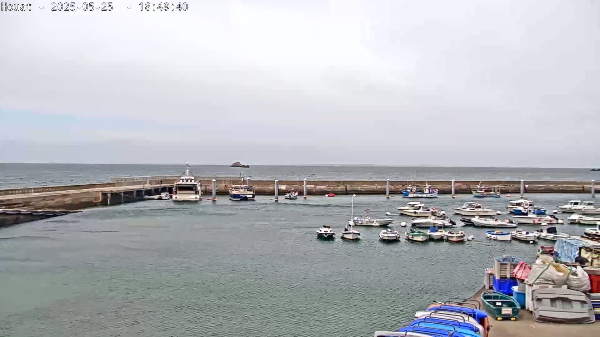 A harbor filled with numerous boats under an overcast sky, with a larger vessel docked near a seawall that extends into a choppy sea.