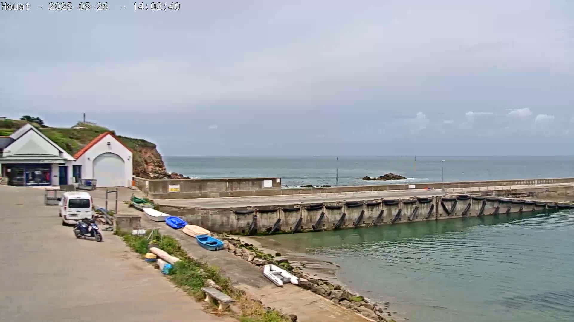 A small harbor with several boats rests beside a building on a rocky coast under a mostly cloudy sky.