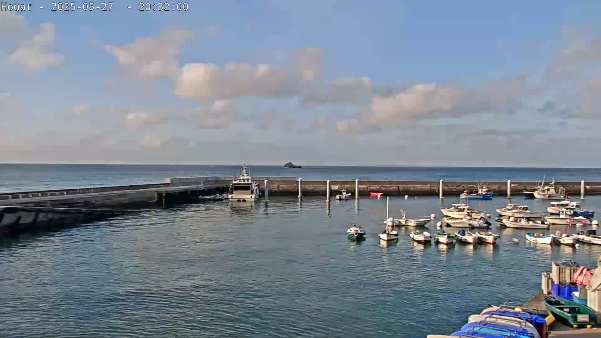 A harbor filled with numerous small boats under a partly cloudy sky, with calm water and a larger vessel docked near a seawall.