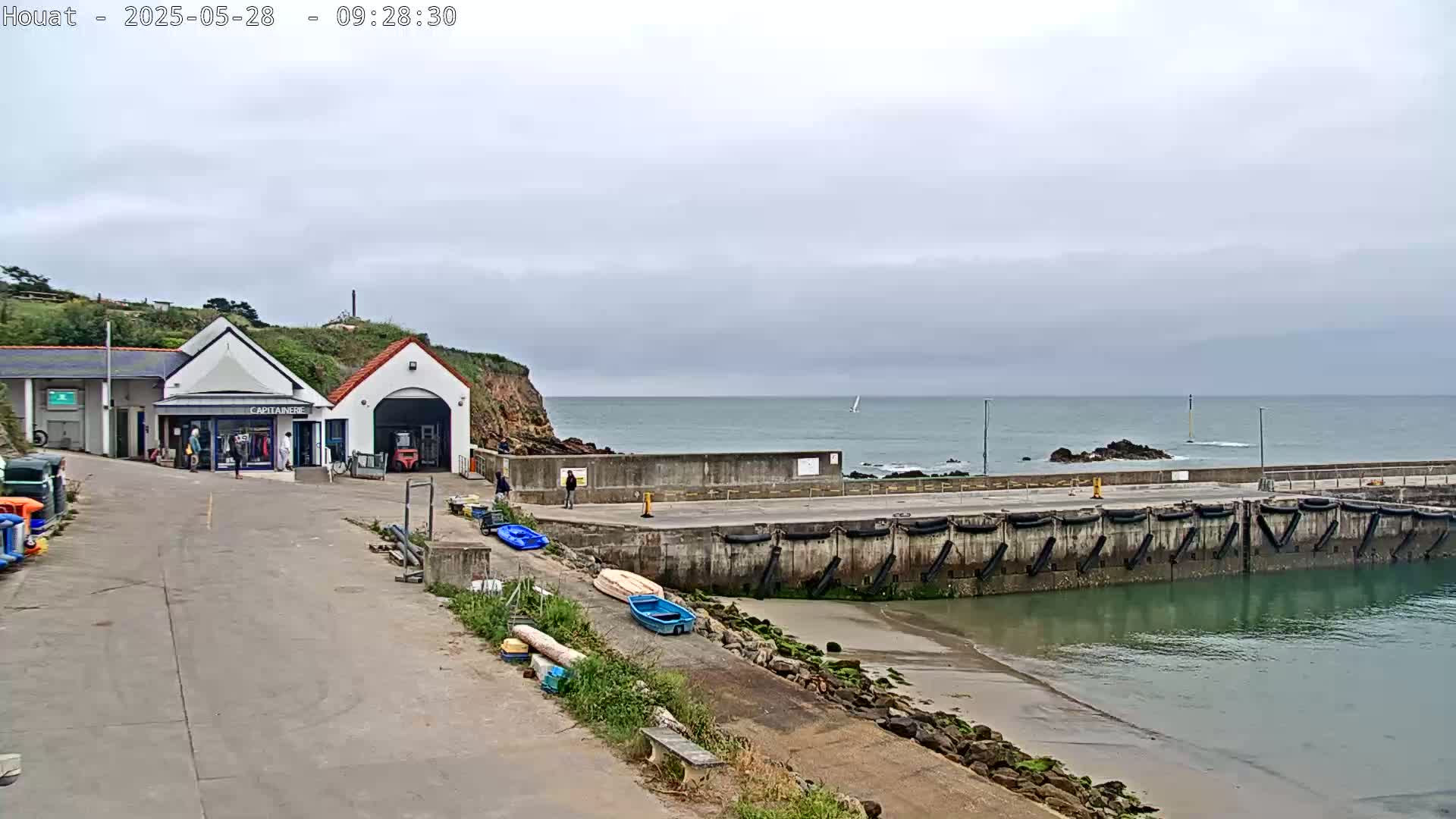 A small harbor with a building and several small boats sits on a calm, overcast day next to a rocky coast and the ocean.