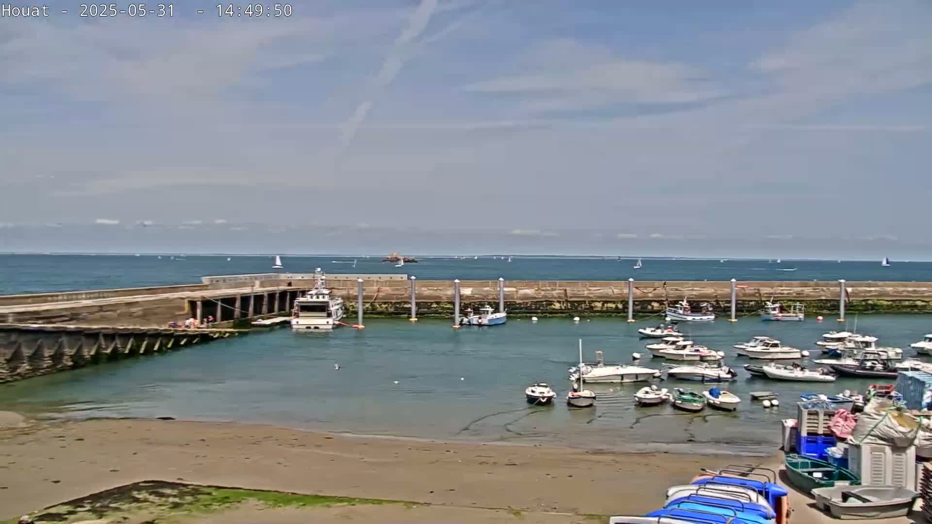 A calm, sunny day shows a harbor filled with numerous boats of varying sizes, nestled within a stone breakwater that extends into a blue ocean dotted with distant sailboats.