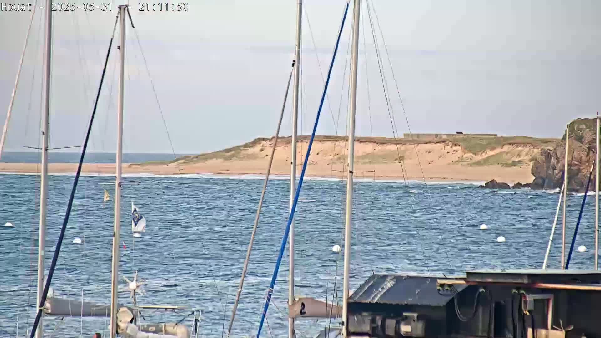 Several sailboats are moored in a harbor with choppy water, and a sandy shore is visible in the background under a mostly clear sky.