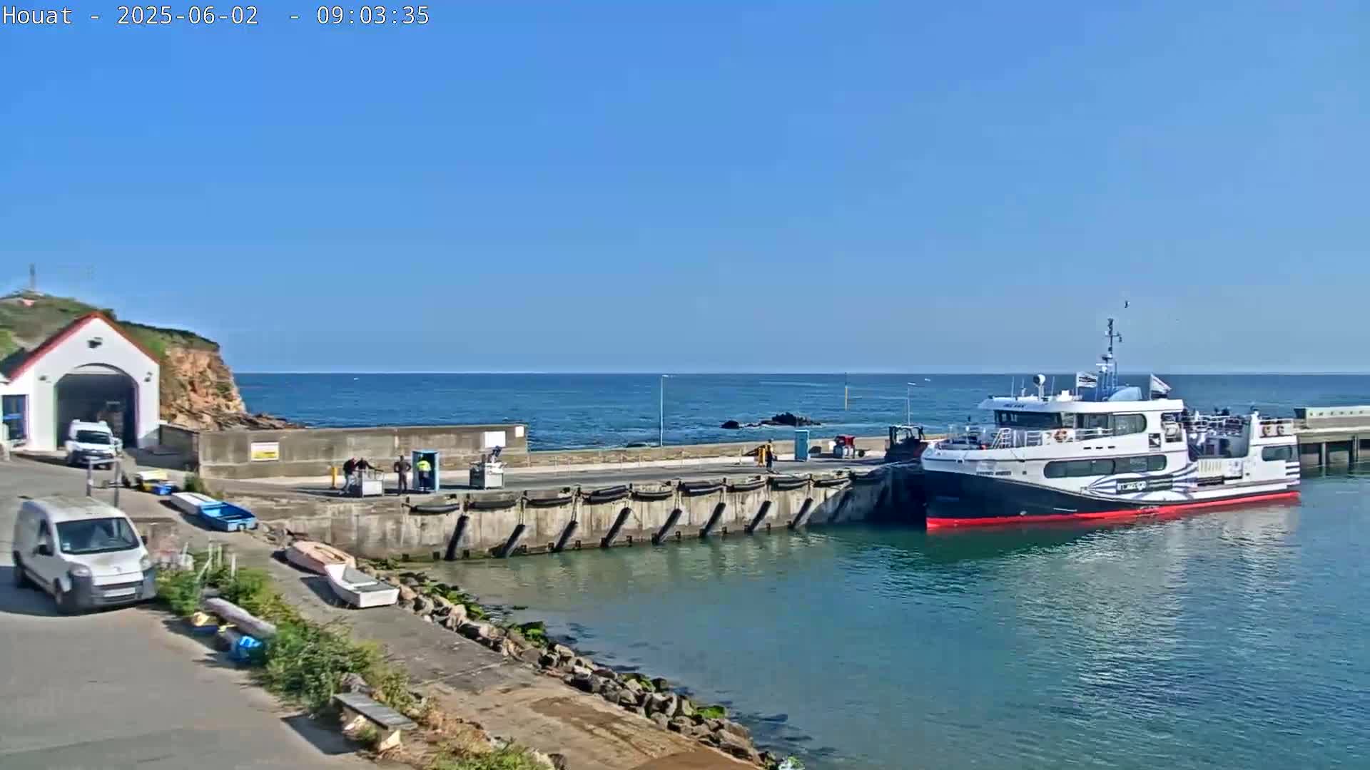 A ferry is docked at a harbor on a sunny day, with a few smaller boats and vehicles nearby.