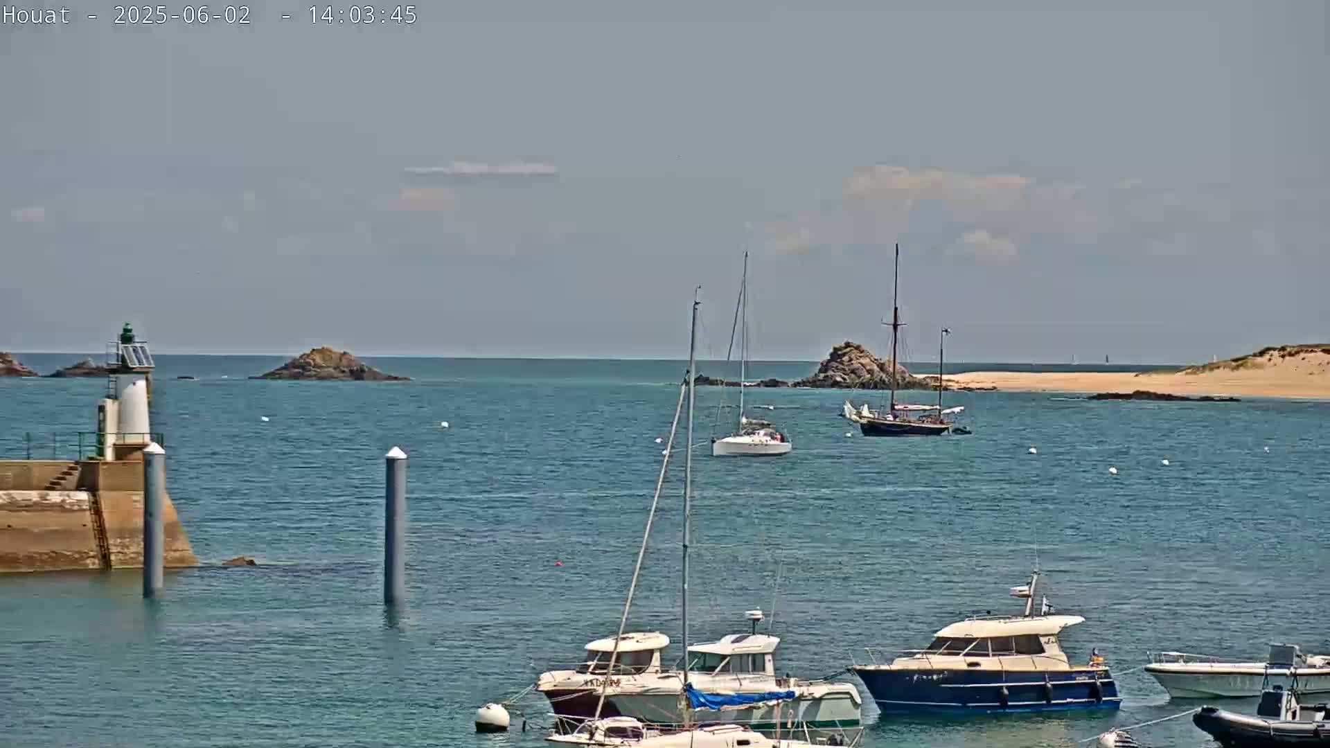 A calm, sunny day shows several sailboats and motorboats in a harbor near a small island and lighthouse.