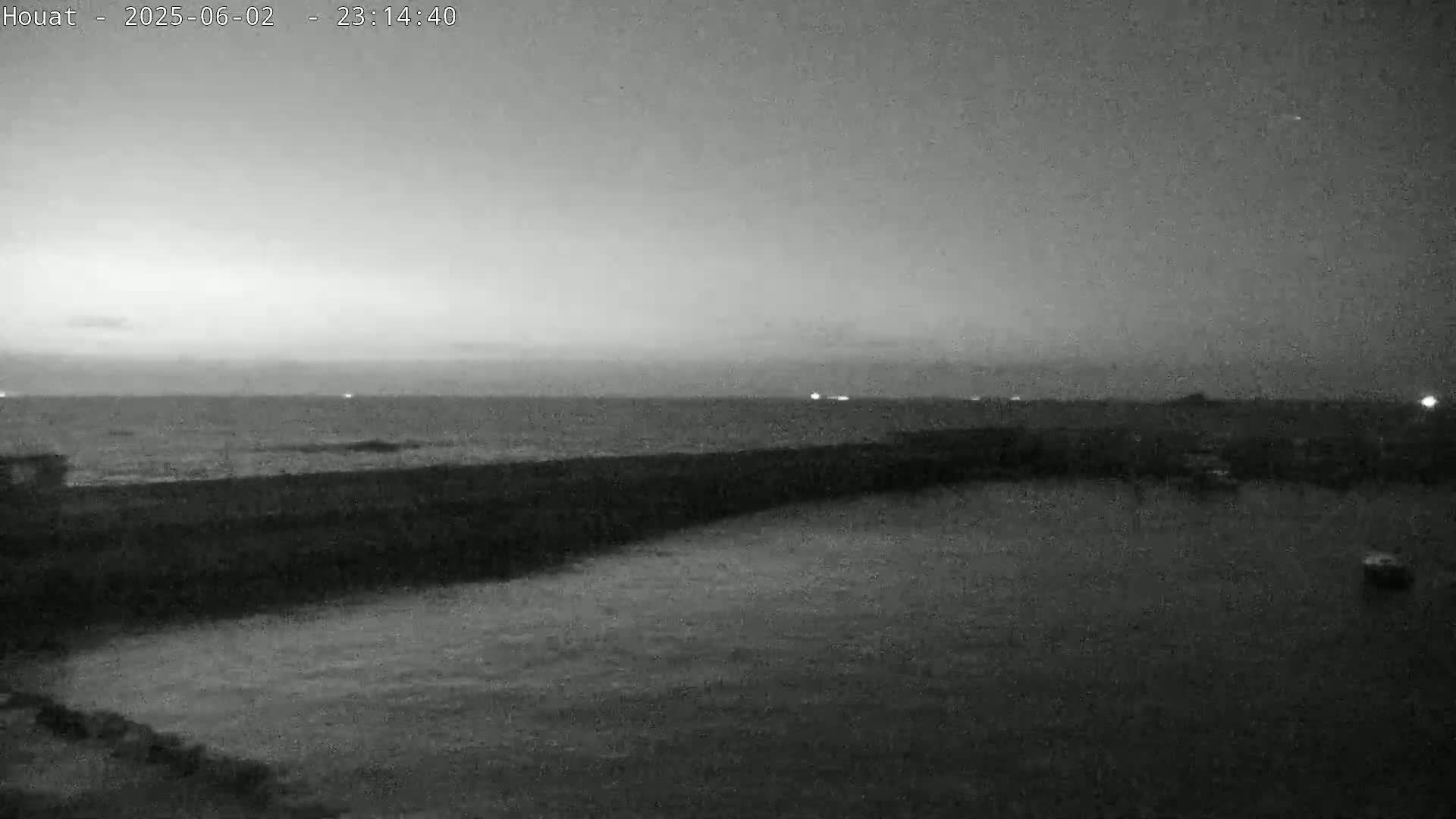 A nighttime grayscale image shows a dark, calm body of water with a dark shoreline and a distant horizon under a cloudy sky.