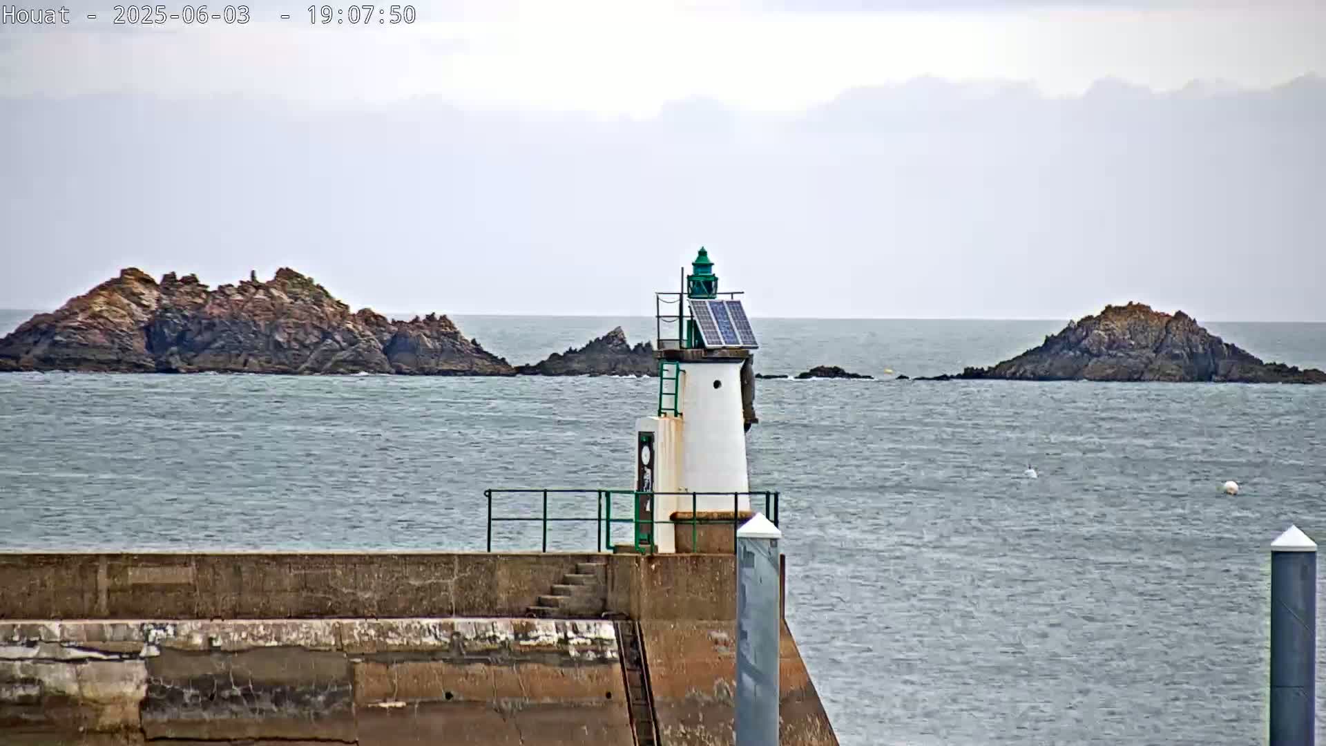 A small, solar-powered lighthouse stands on a concrete pier overlooking a calm sea with two rocky islands in the distance under an overcast sky.