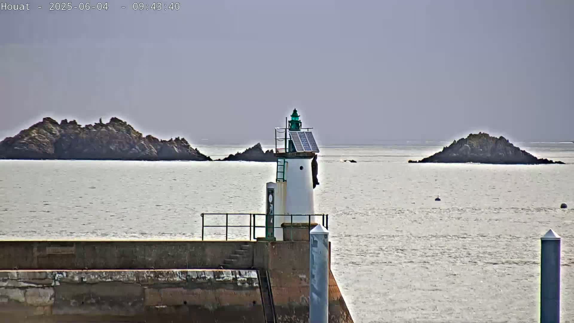 A small lighthouse with a solar panel stands on a stone jetty overlooking calm, gray water and two small rocky islands under a hazy sky.