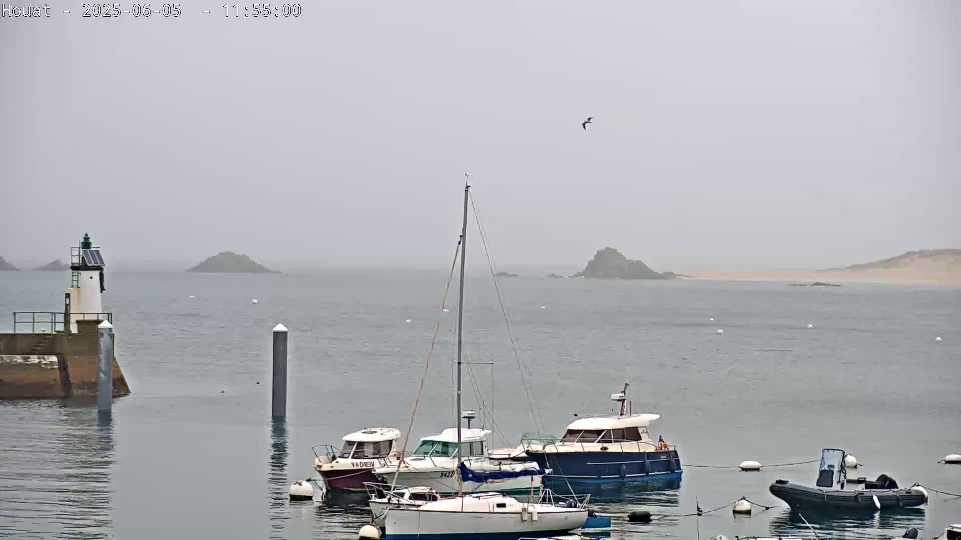 A hazy, overcast day shows several sailboats and motorboats moored in a harbor near a small lighthouse and distant islands.