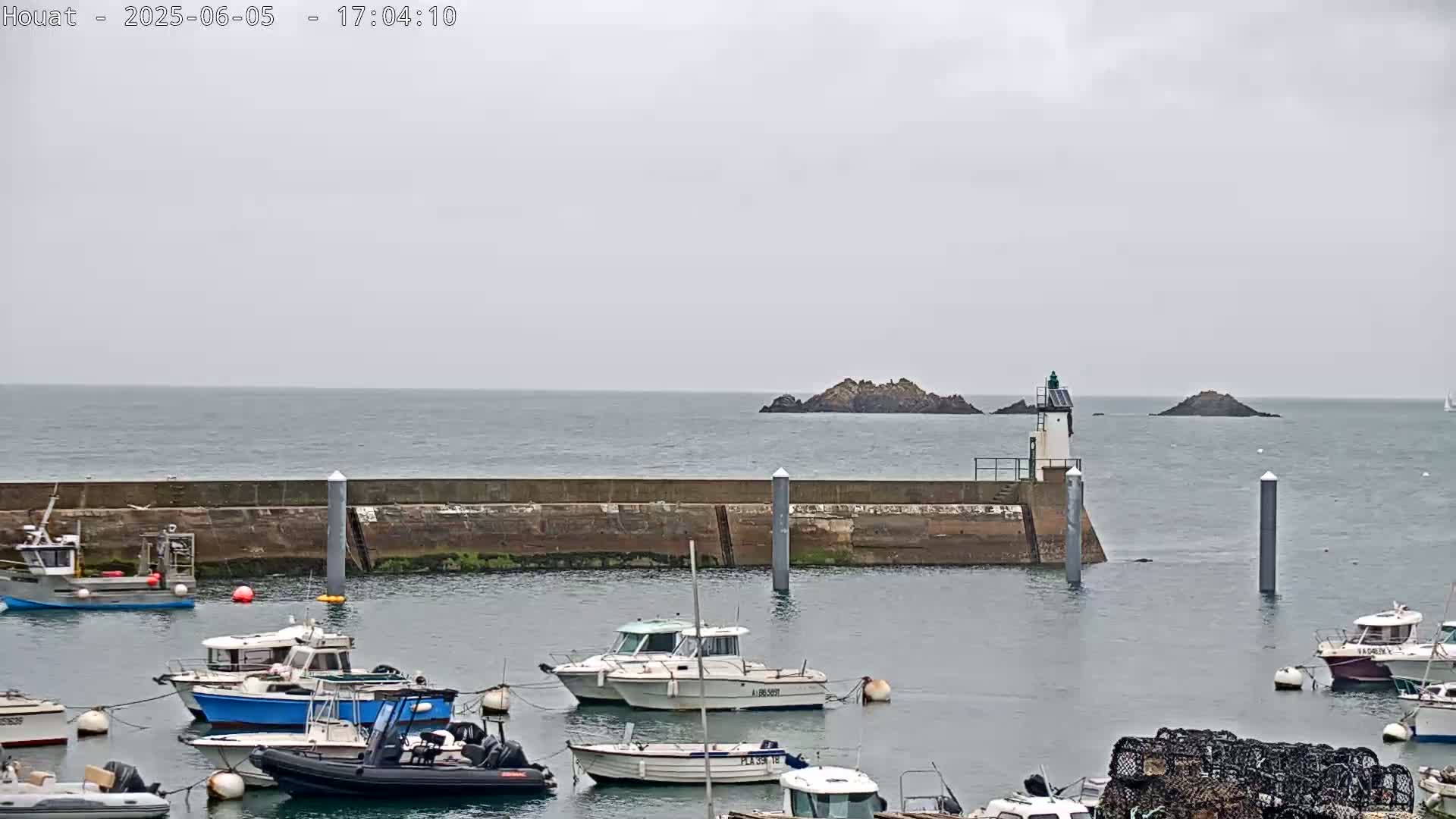 A harbor filled with several boats is seen under an overcast sky, with a small island and lighthouse visible in the distance.