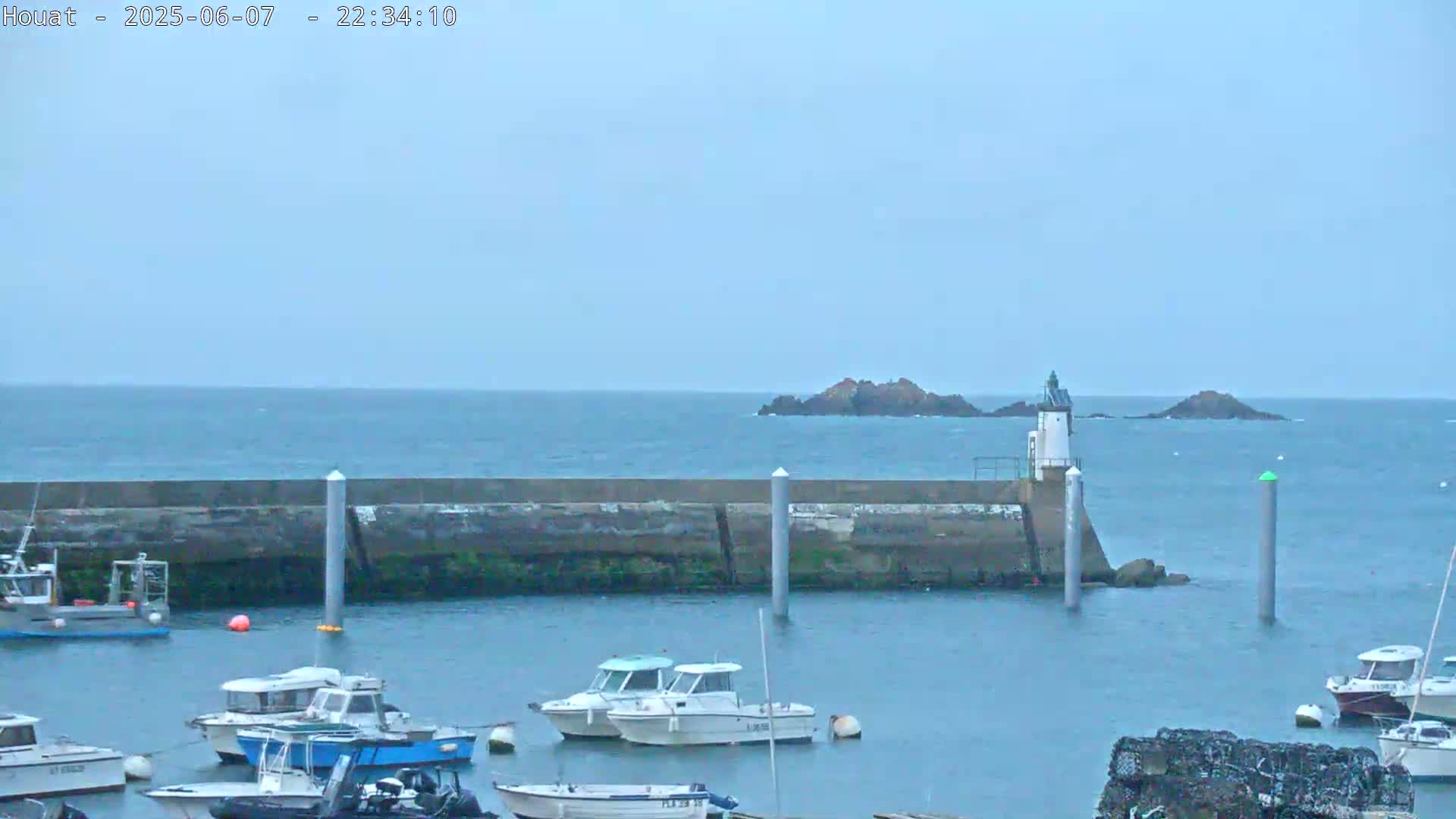 A harbor filled with small boats is partially protected by a seawall, beyond which a small lighthouse and rocky islands are visible under an overcast sky.