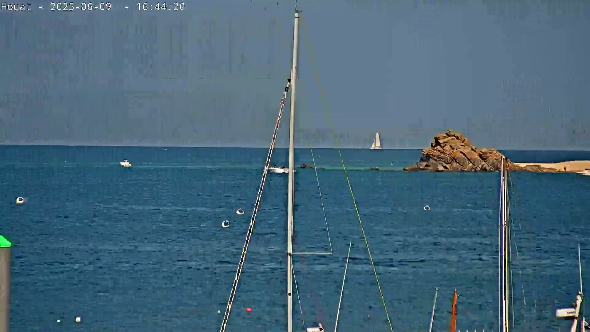 A calm, sunny day at sea shows several sailboats and a rocky outcropping, with a sailboat in the distance.