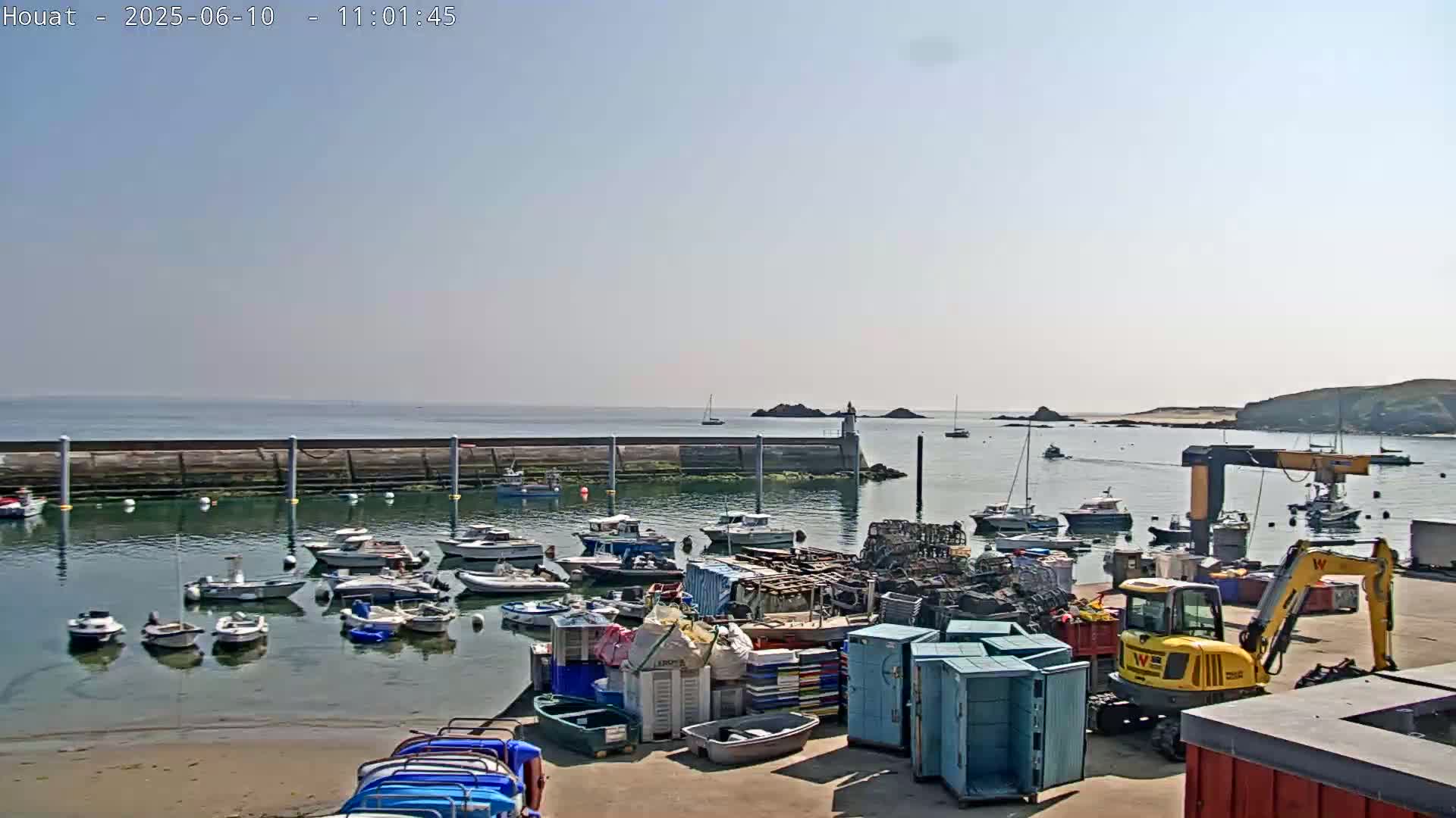 A harbor filled with numerous small boats is shown on a sunny day, with a small yellow excavator and various storage containers on the dock.