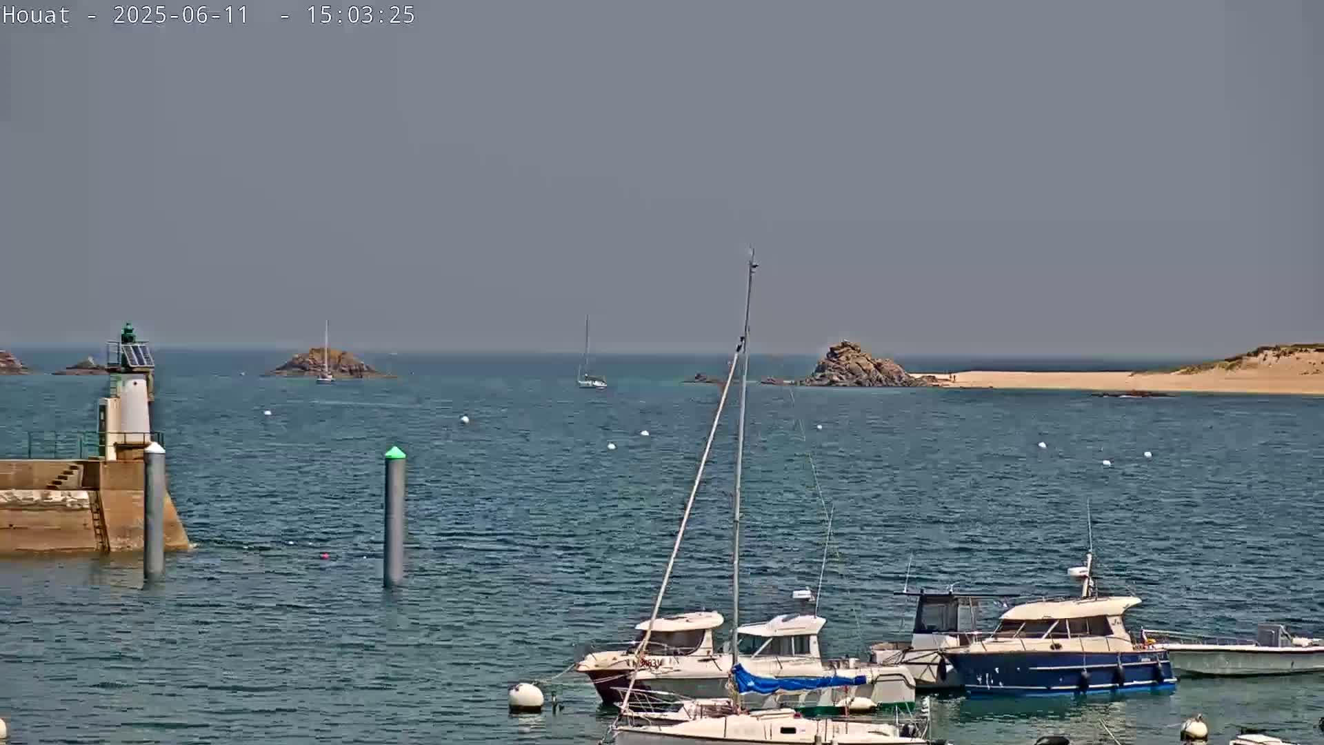 A harbor scene on a sunny day shows several boats moored near a lighthouse and a rocky shoreline with a sandy beach in the distance.