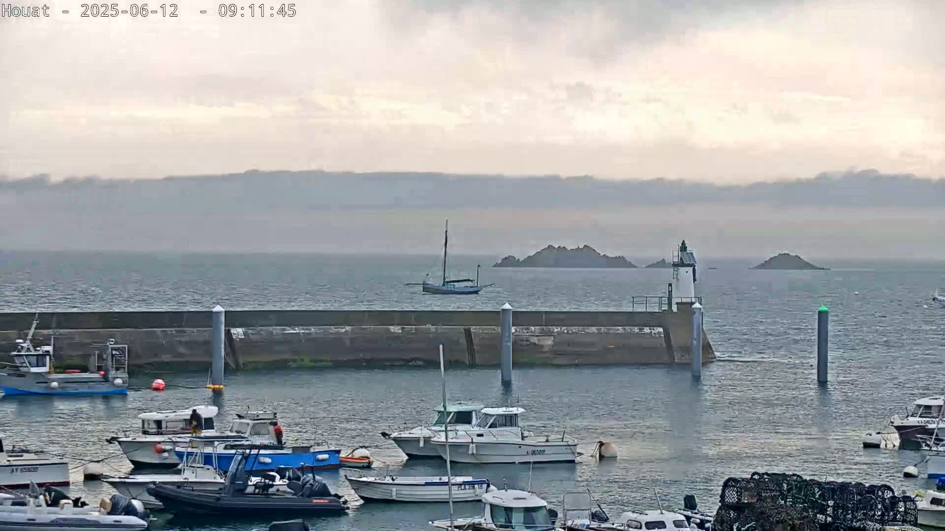 A harbor filled with numerous boats is shown under an overcast sky, with a sailboat and a lighthouse visible in the distance across calm water.