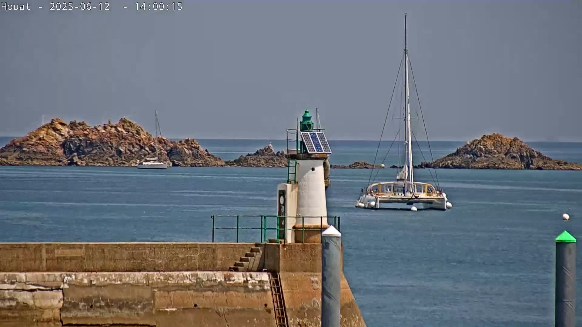A small lighthouse with a solar panel sits on a stone jetty overlooking a calm sea with rocky islands and a sailboat in the distance under a clear sky.