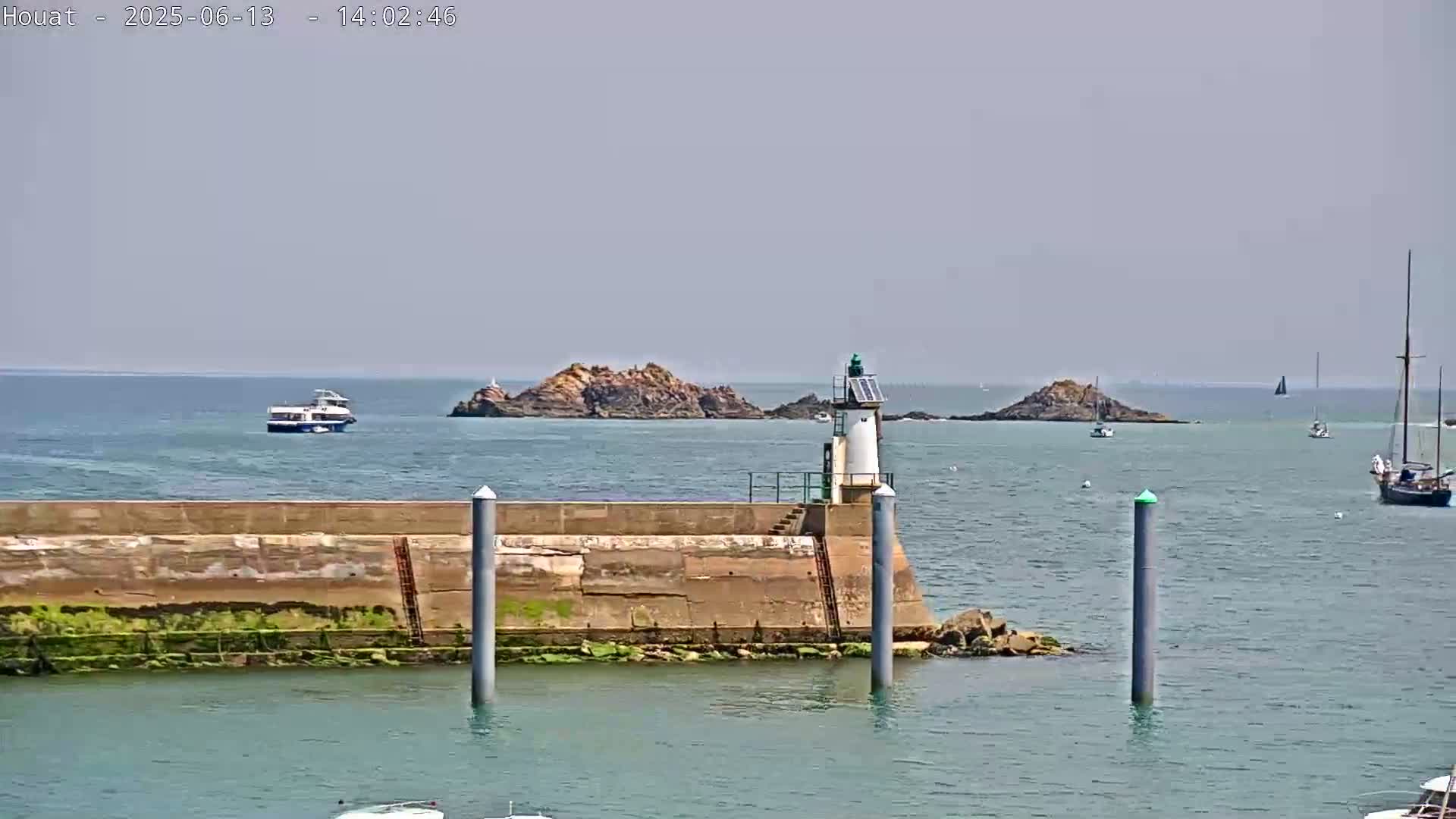 A small lighthouse sits on a seawall next to calm, blue water, with a rocky island, a ferry, and several sailboats visible in the distance under a hazy sky.