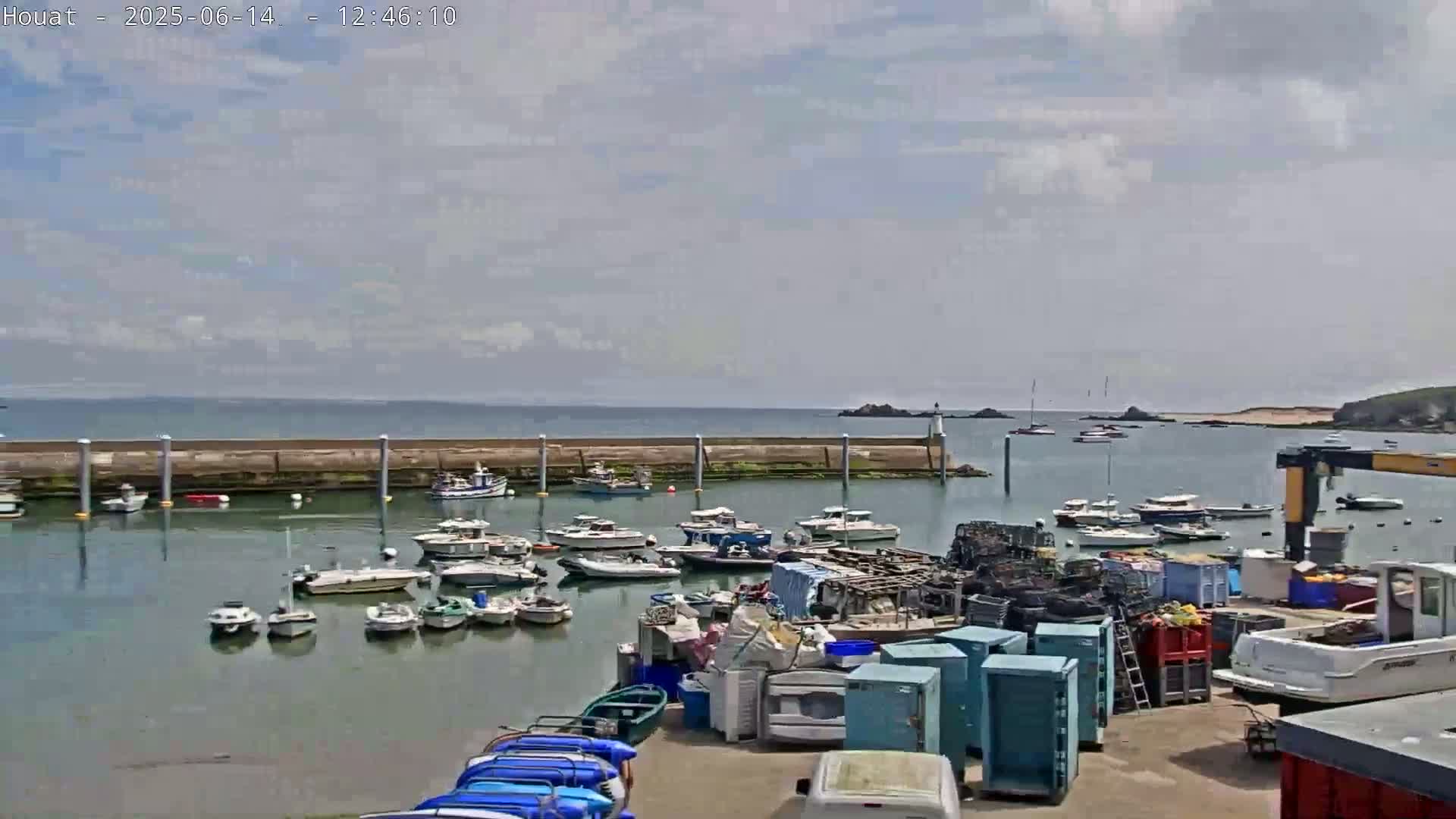 A harbor filled with numerous boats is seen under a partly cloudy sky, with a calm sea extending to a distant shoreline.