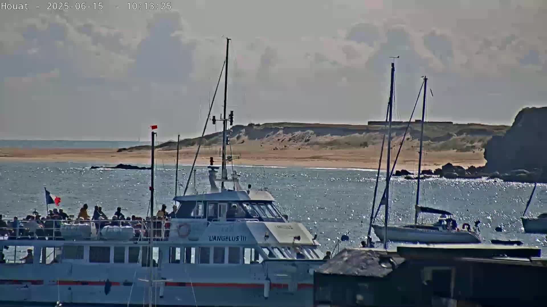 A passenger ferry, named "Langueux III," is in the water near a sandy shore under partly cloudy skies, with sailboats moored nearby.