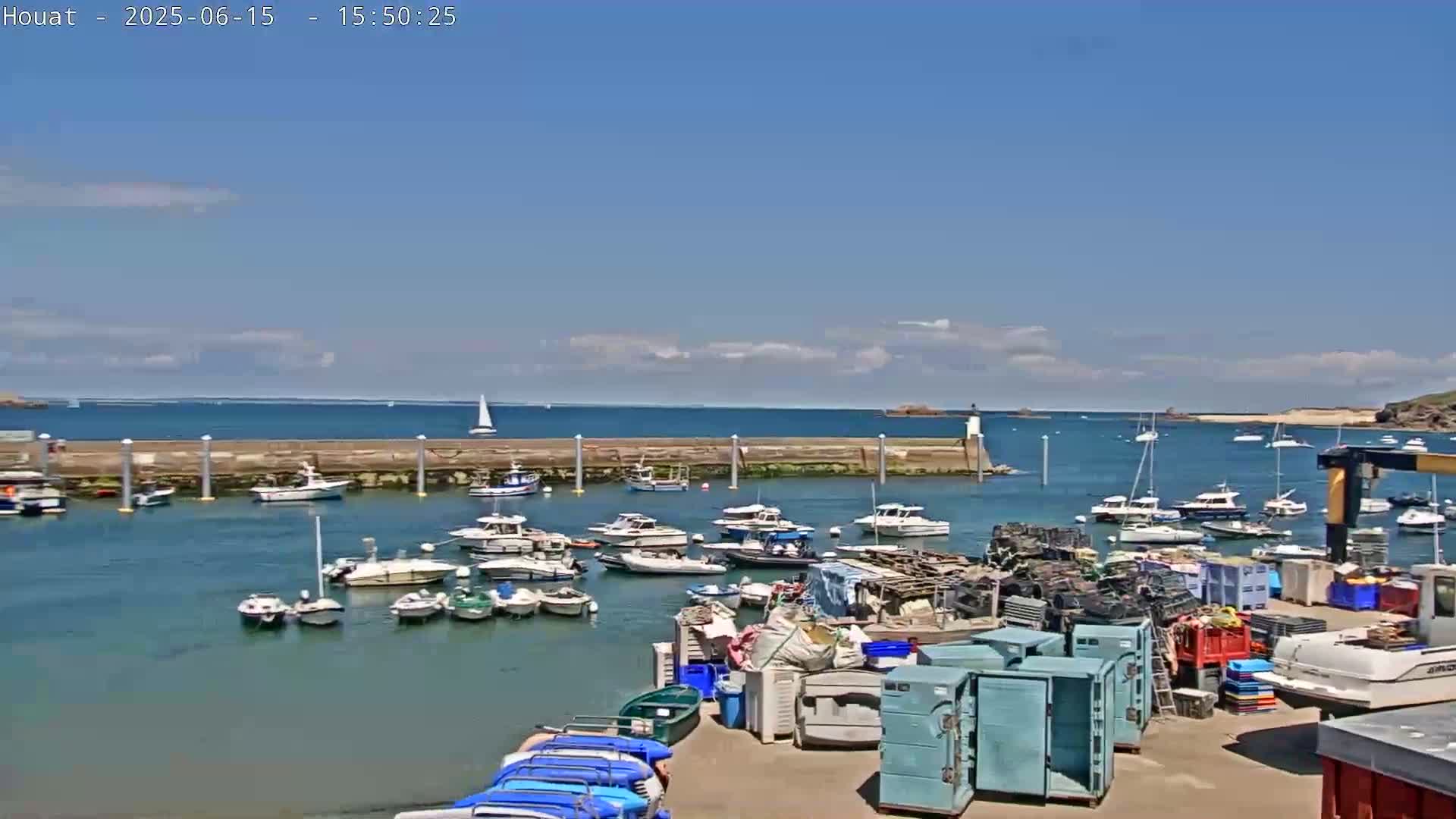 A sunny day shows numerous boats moored in a harbor, with additional boats at anchor in the calm waters beyond a stone seawall.