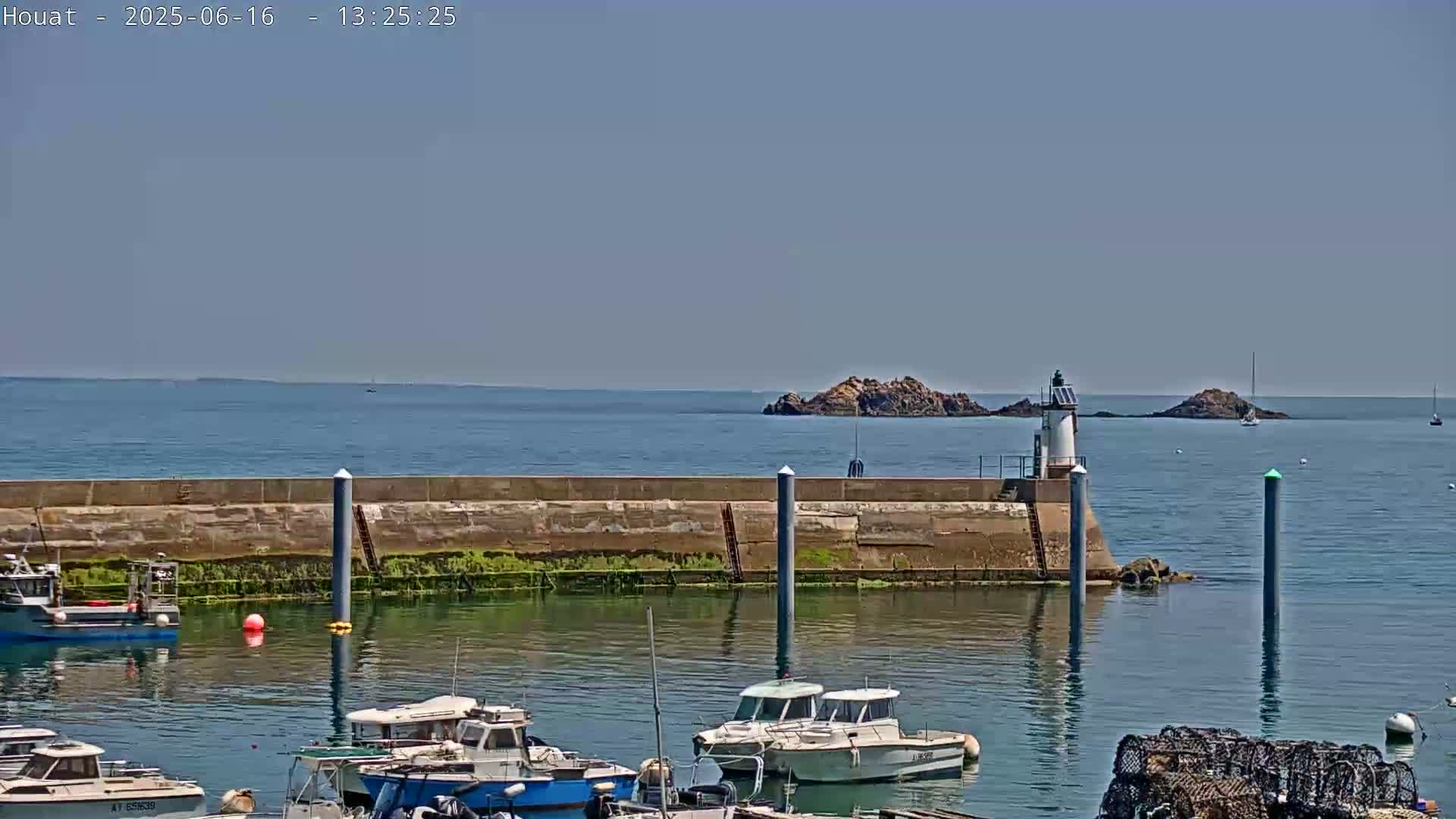 A harbor filled with several boats sits behind a seawall, with a small lighthouse and rocky islands visible in the calm ocean under a clear blue sky.