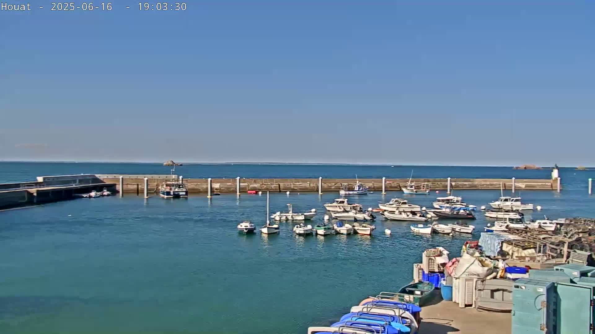 A calm harbor filled with numerous boats is enclosed by a seawall under a clear blue sky.