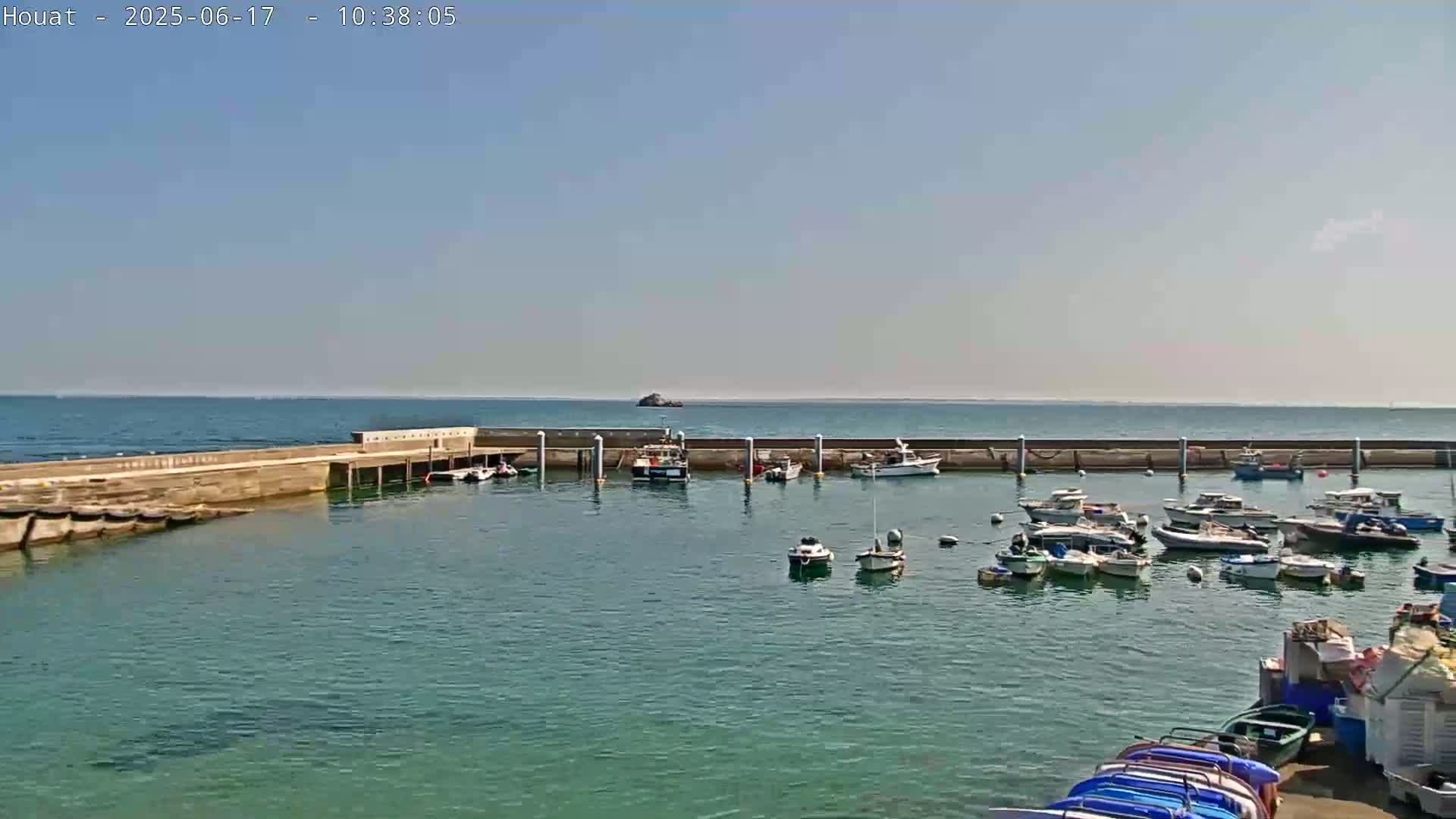 A calm, sunny day shows a small harbor filled with numerous boats, enclosed by a concrete seawall, with a distant island visible on the horizon.