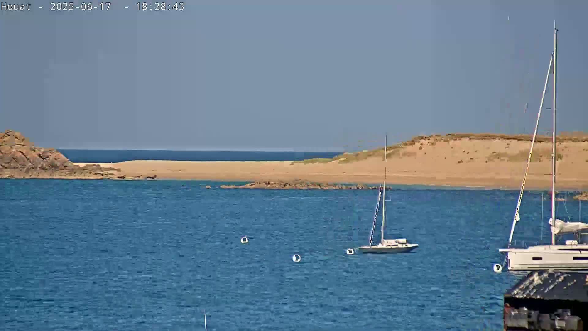 A calm, sunny day shows a sandy beach and rocky shoreline with two sailboats anchored in the clear blue water.