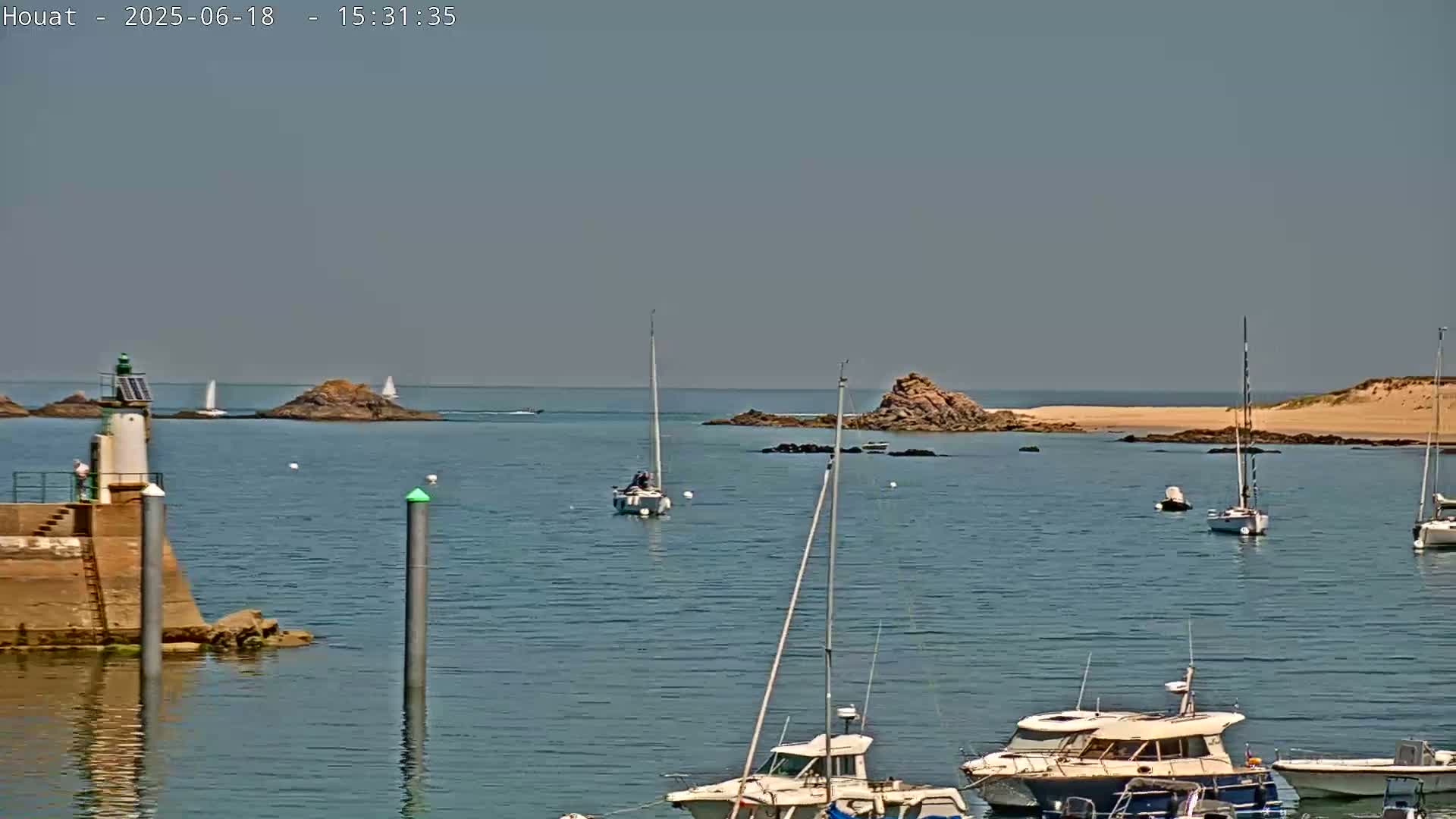 A calm, sunny day shows a harbor with several sailboats and motorboats, a small lighthouse, and rocky islands in the distance.