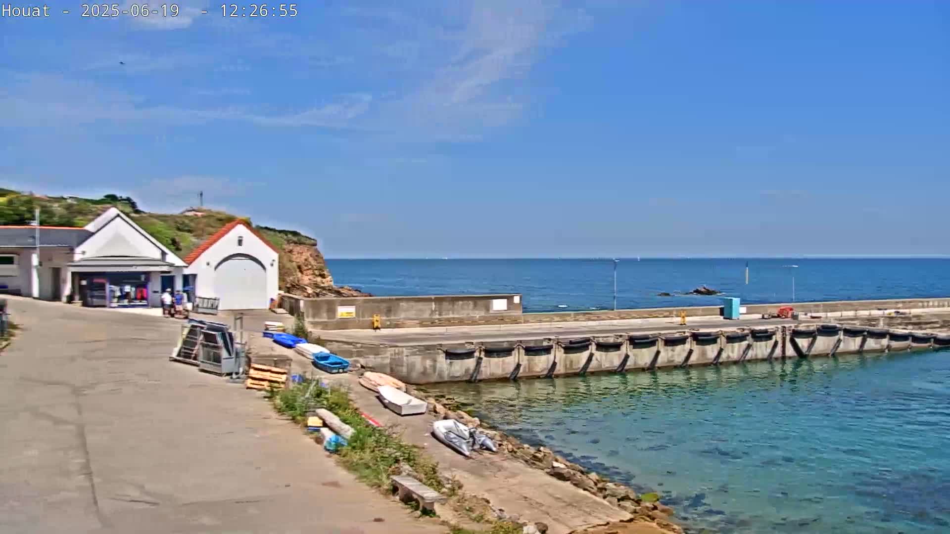 A small harbor with a building and several boats sits on a rocky coast under a clear blue sky.