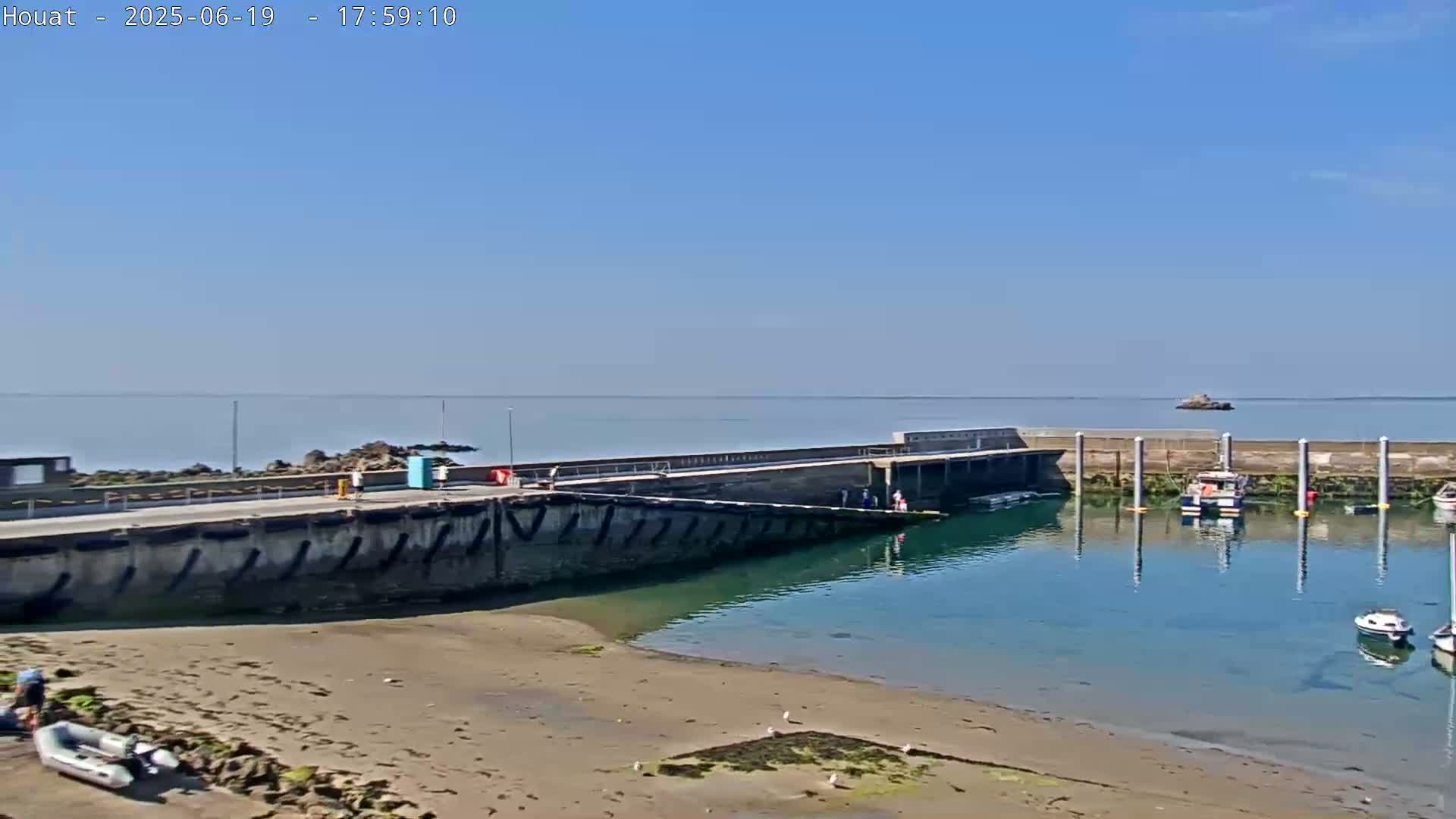 A calm, sunny day shows a small harbor with several boats moored, a concrete pier, and a sandy beach.
