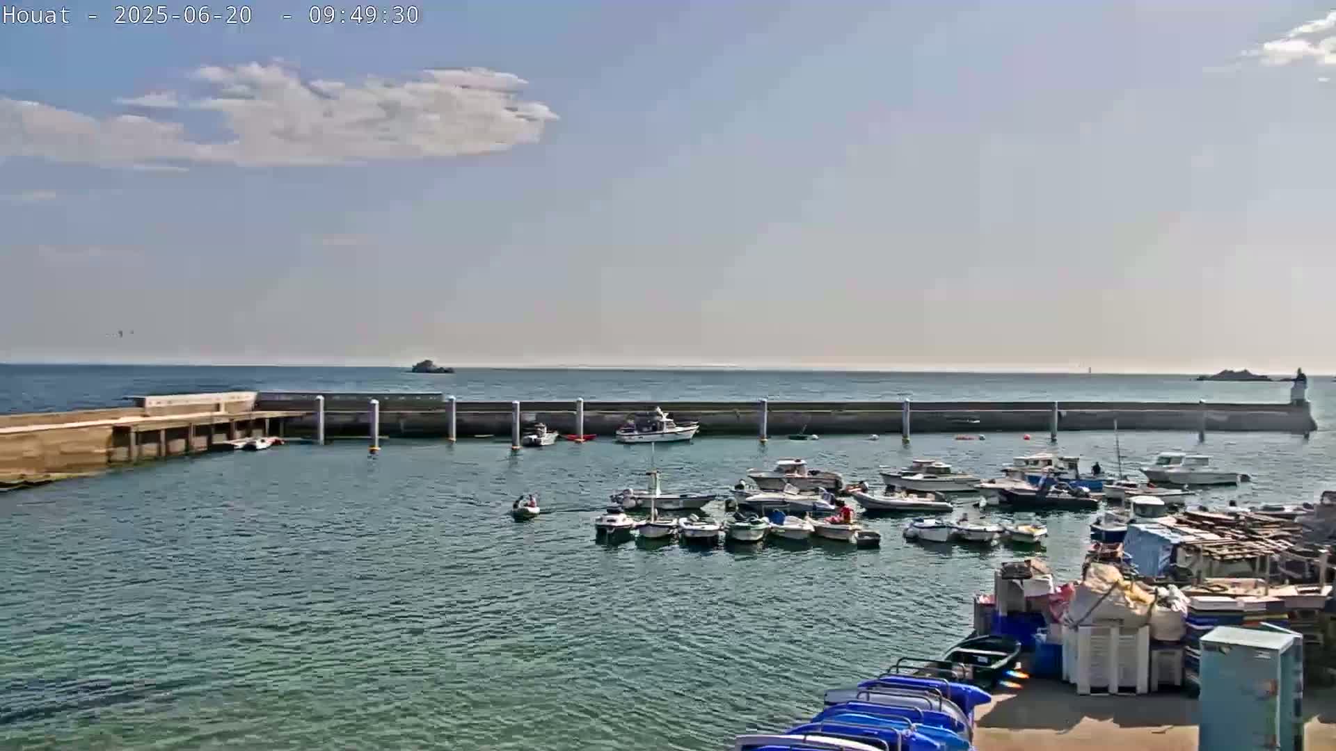 A harbor filled with numerous small boats is nestled inside a stone breakwater on a partly cloudy day.