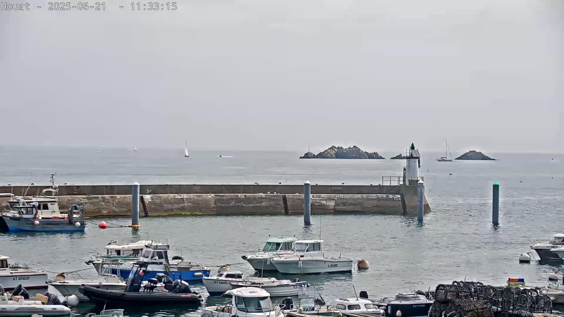 A harbor filled with numerous boats is situated before a calm sea with a lighthouse and small islands in the distance under an overcast sky.
