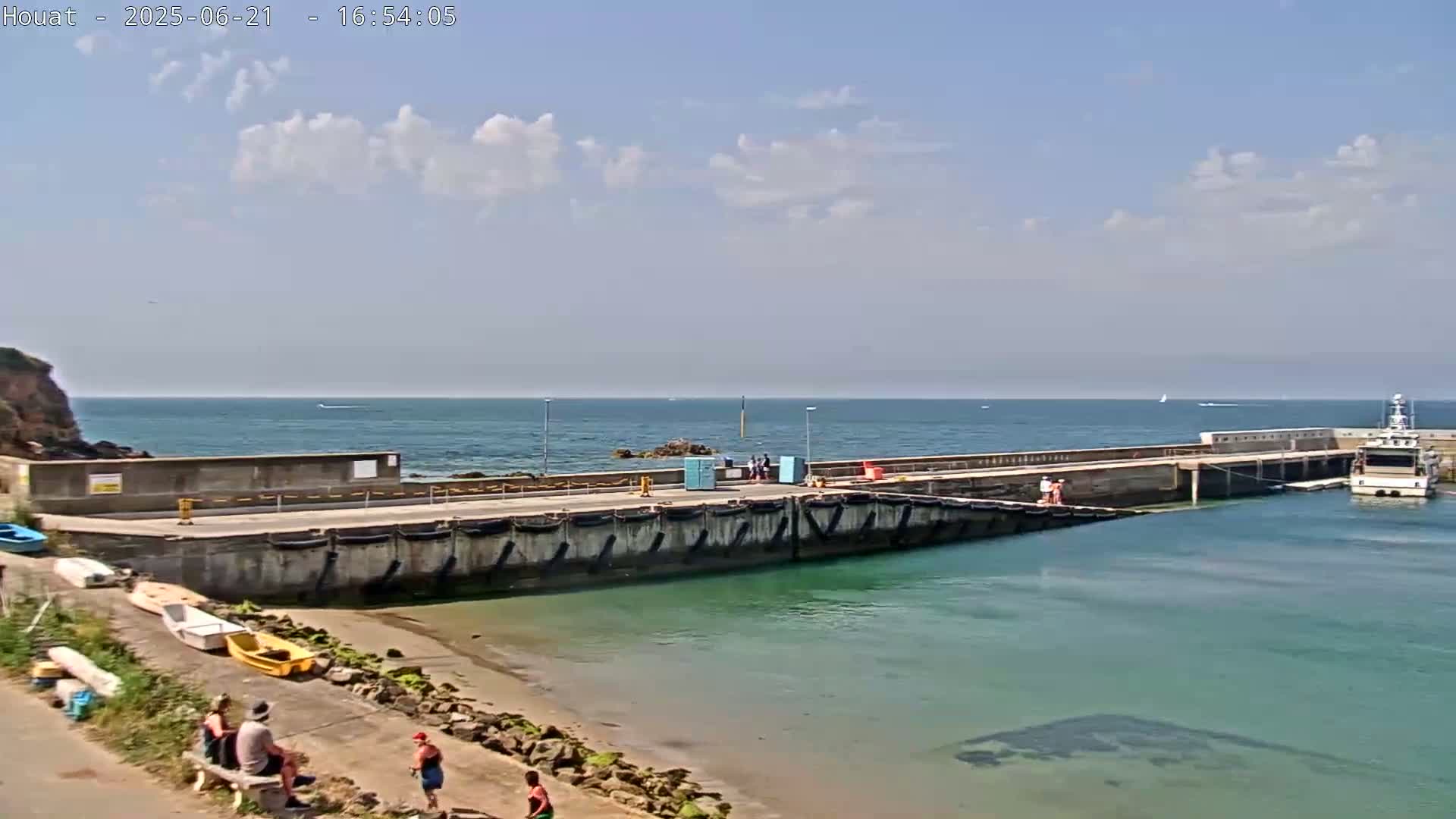 A concrete pier extends into a calm, blue ocean under a partly cloudy sky, with a small sandy beach and several people visible nearby.
