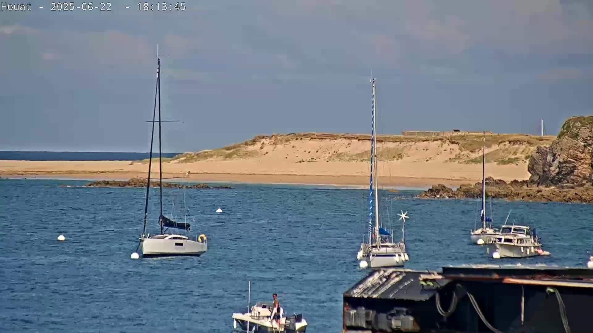 Several sailboats and motorboats are anchored in calm waters near a sandy beach and rocky shoreline under a mostly clear sky.