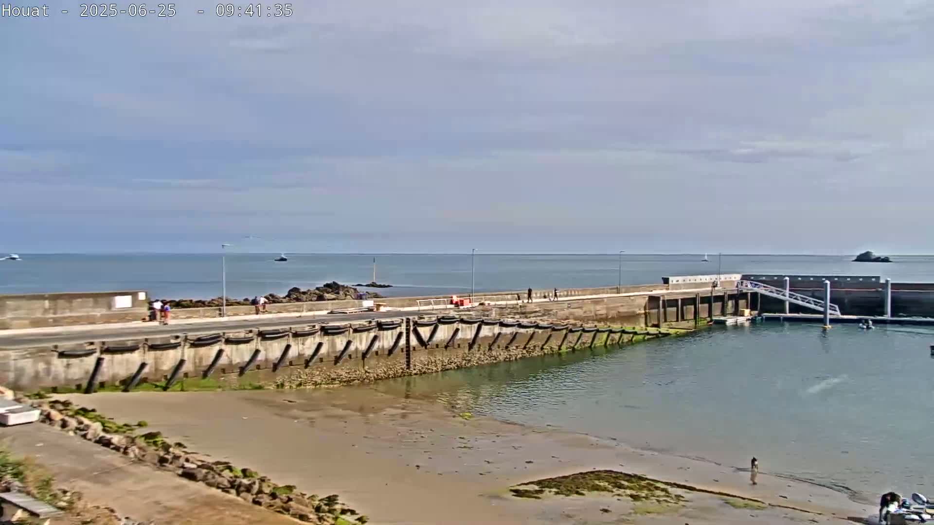 A mostly sunny day reveals a small harbor with a concrete wall, a ramp for boats, and several people walking along the seawall overlooking calm water.