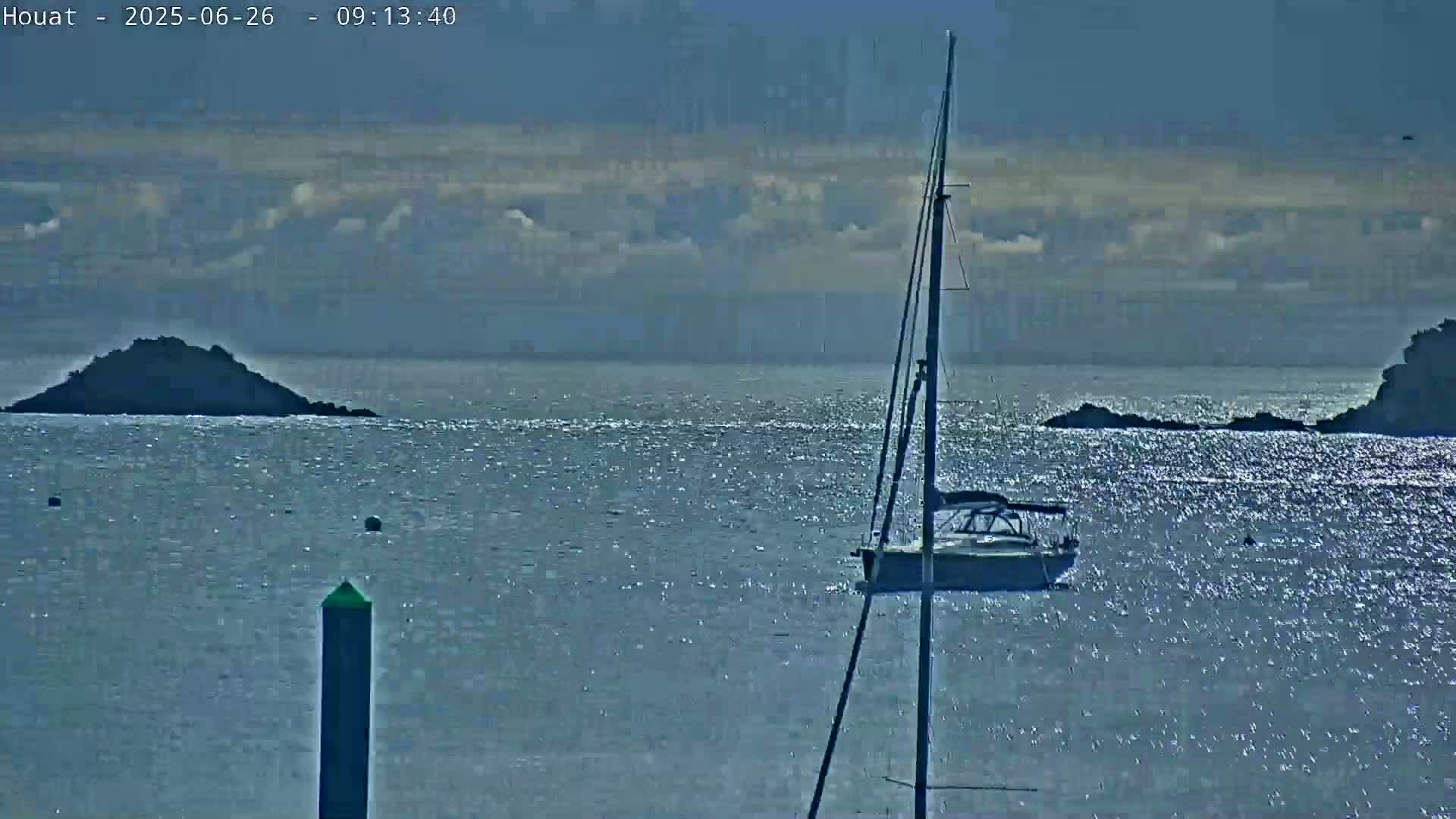 A sailboat is anchored in a calm, sparkling body of water near several small, dark islands under a partly cloudy sky.