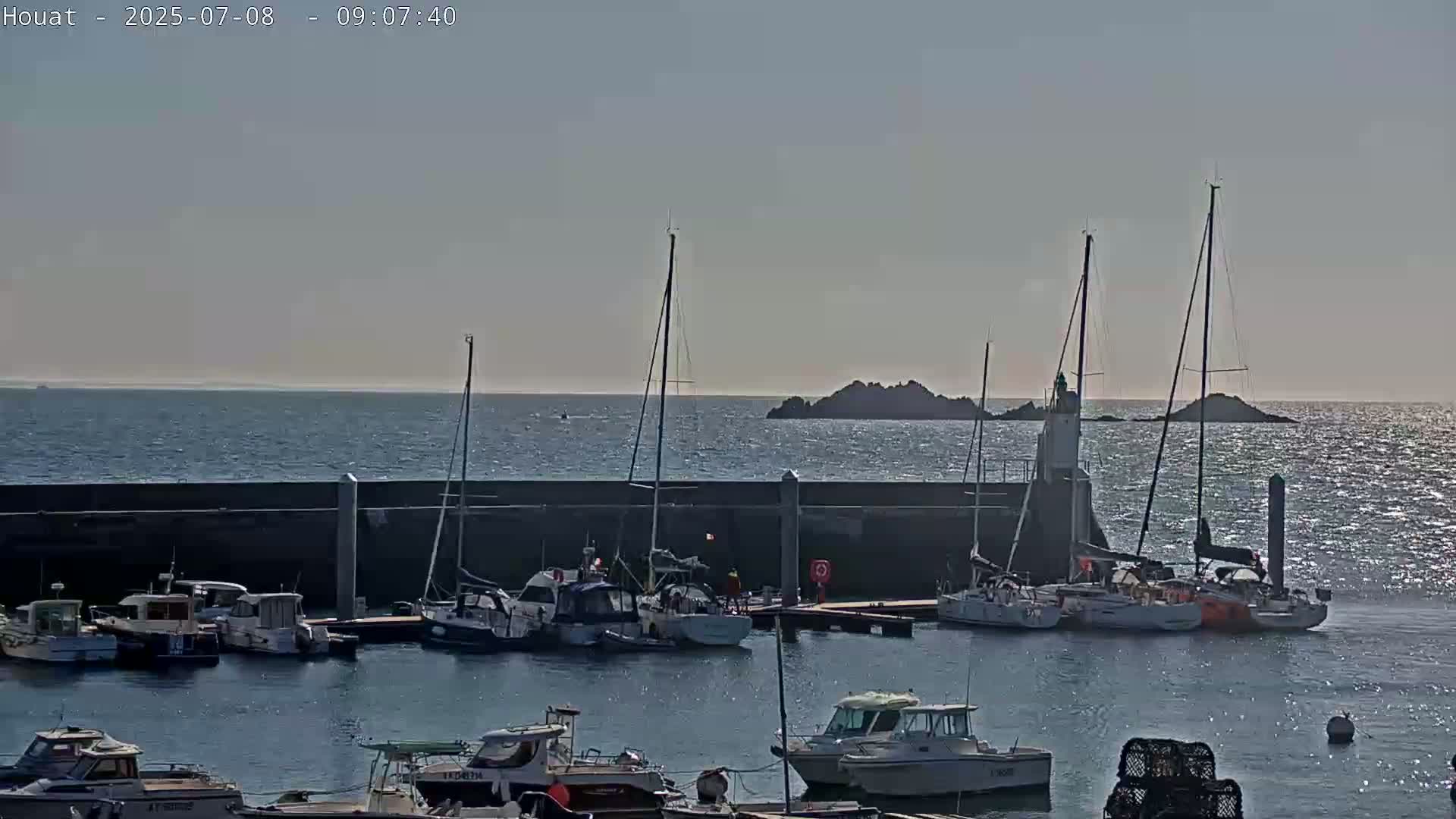 Several sailboats and motorboats are moored in a harbor next to a low stone wall, with rocky islands and a lighthouse visible in the calm, sunlit sea beyond.